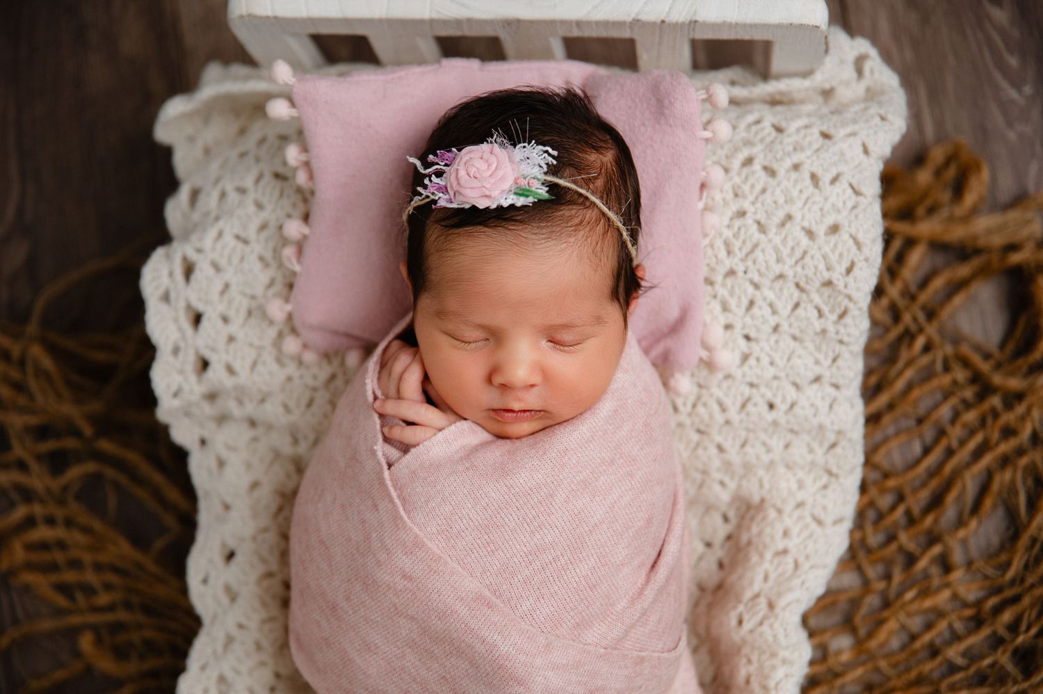Peaceful newborn wrapped in pink swaddle sleeping on white blanket with floral headband.