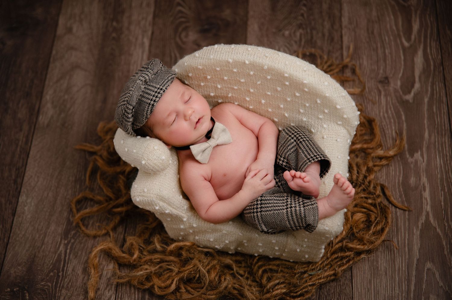 Sleeping newborn in knit cap and outfit curled up in white-lined basket on wooden floor.