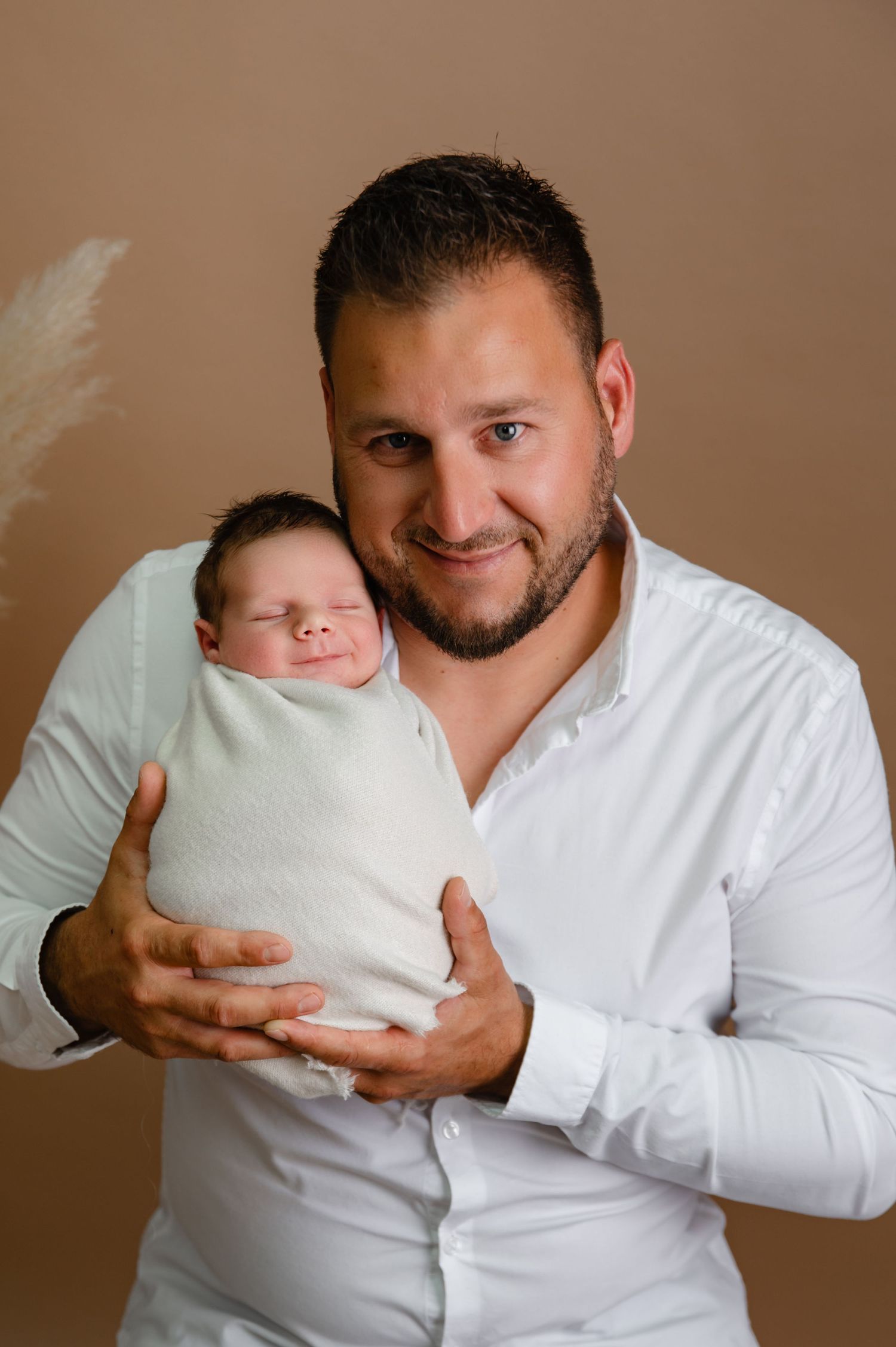 Father in white shirt holding swaddled newborn against warm brown backdrop.