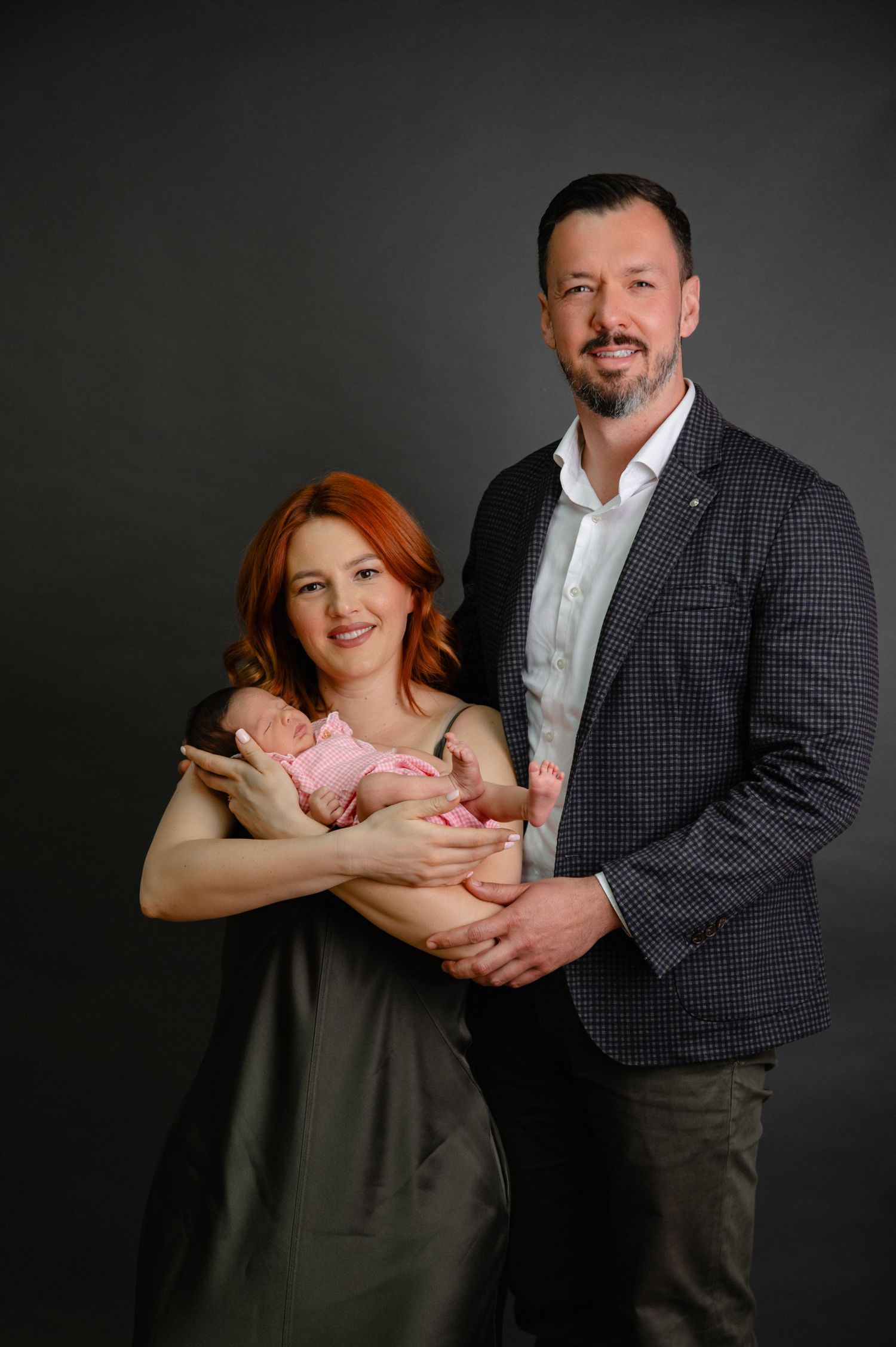 Professional studio portrait of couple holding newborn baby against dark background.