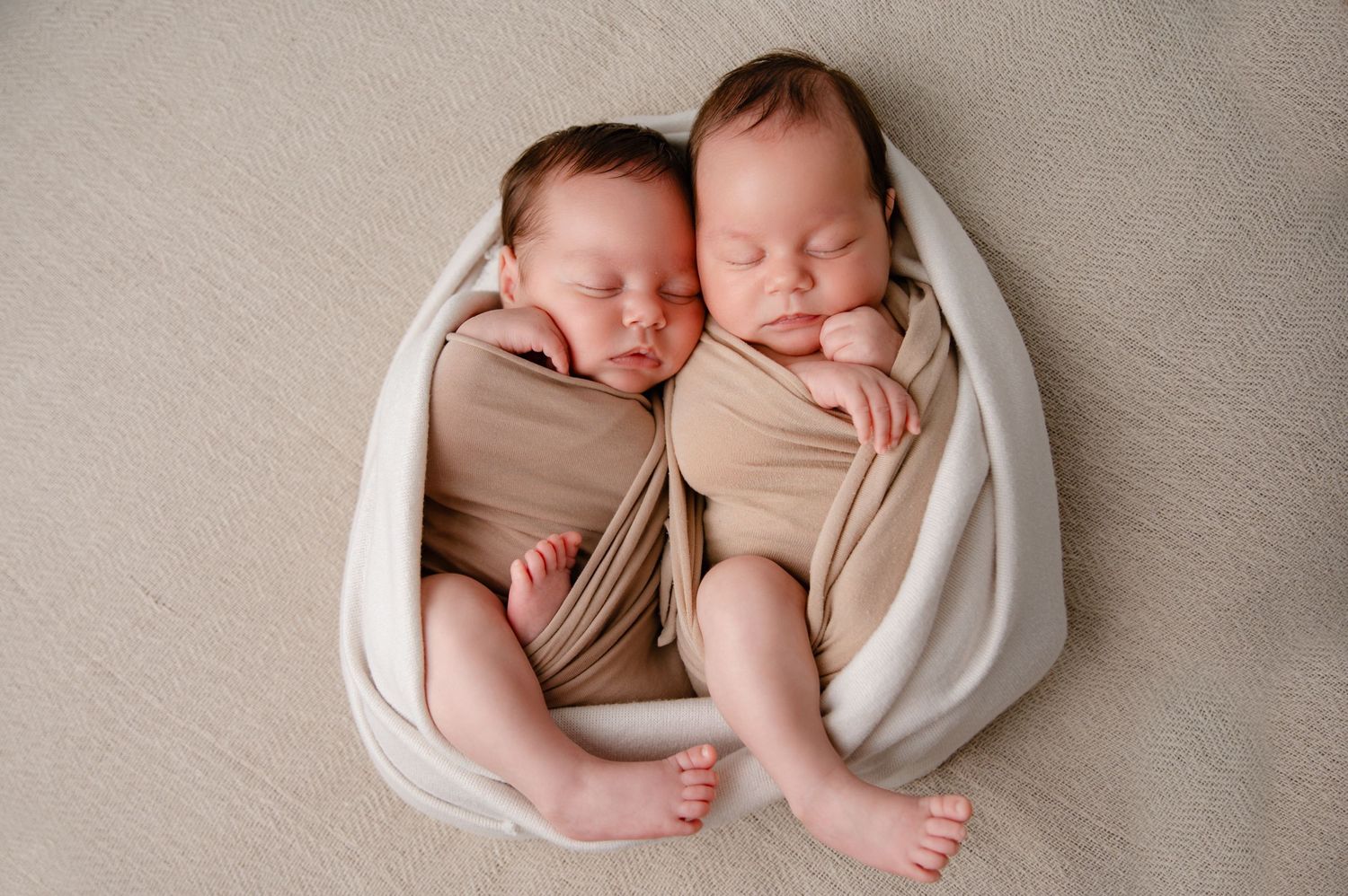 Two newborn twins sleeping peacefully wrapped in beige and white fabric.