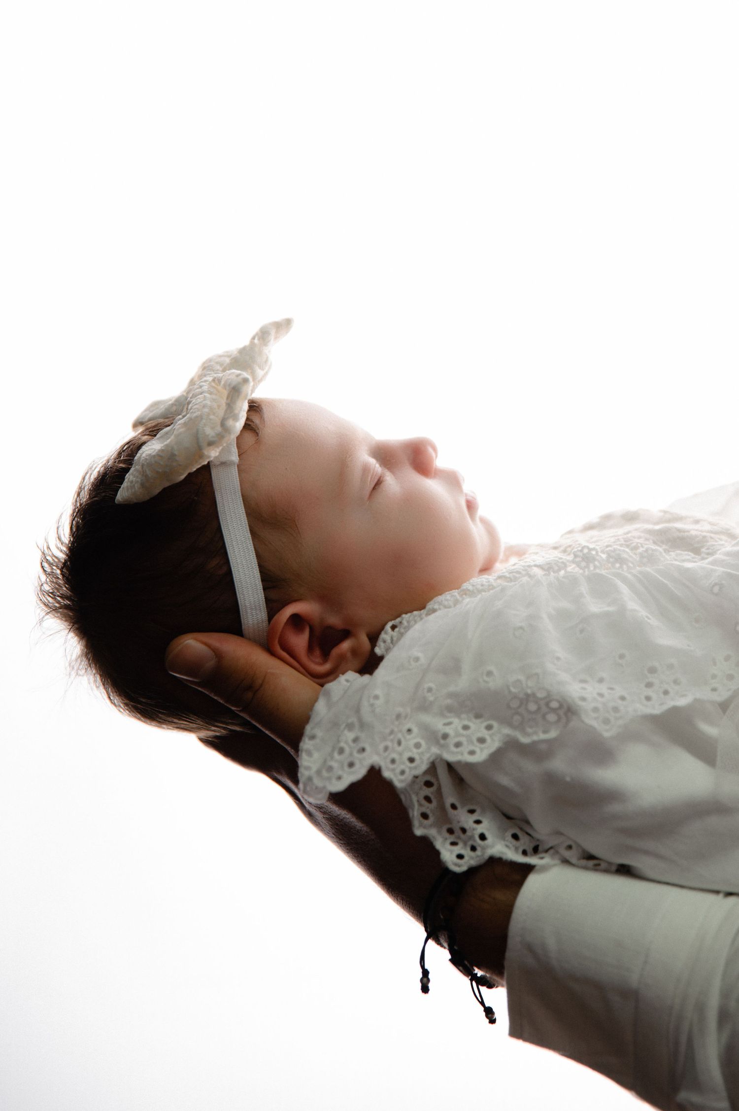 Close-up profile of a sleeping baby wearing a white lace dress and delicate bow headband.