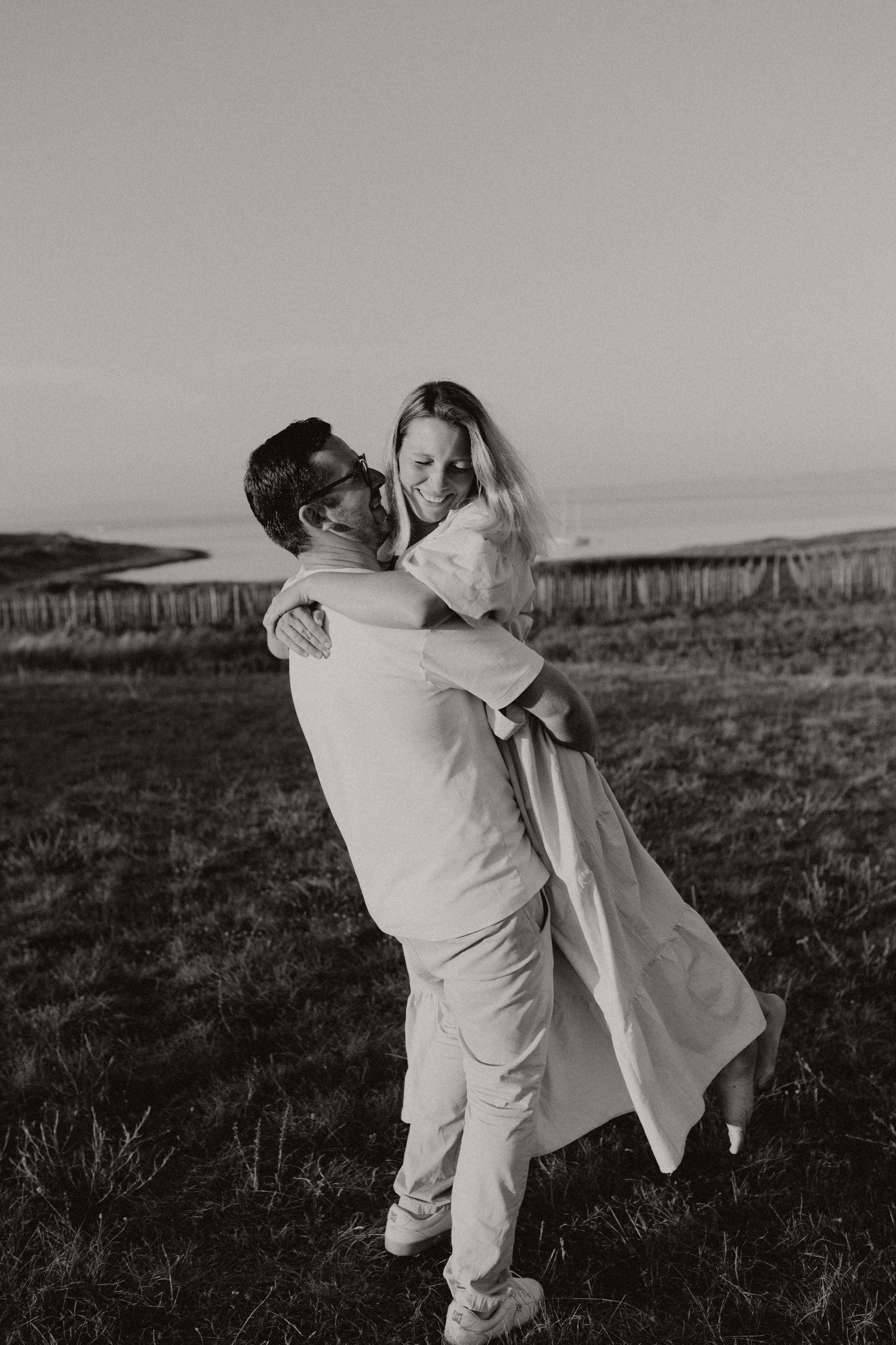 A romantic black and white photo of a couple embracing in a grassy field at sunset.