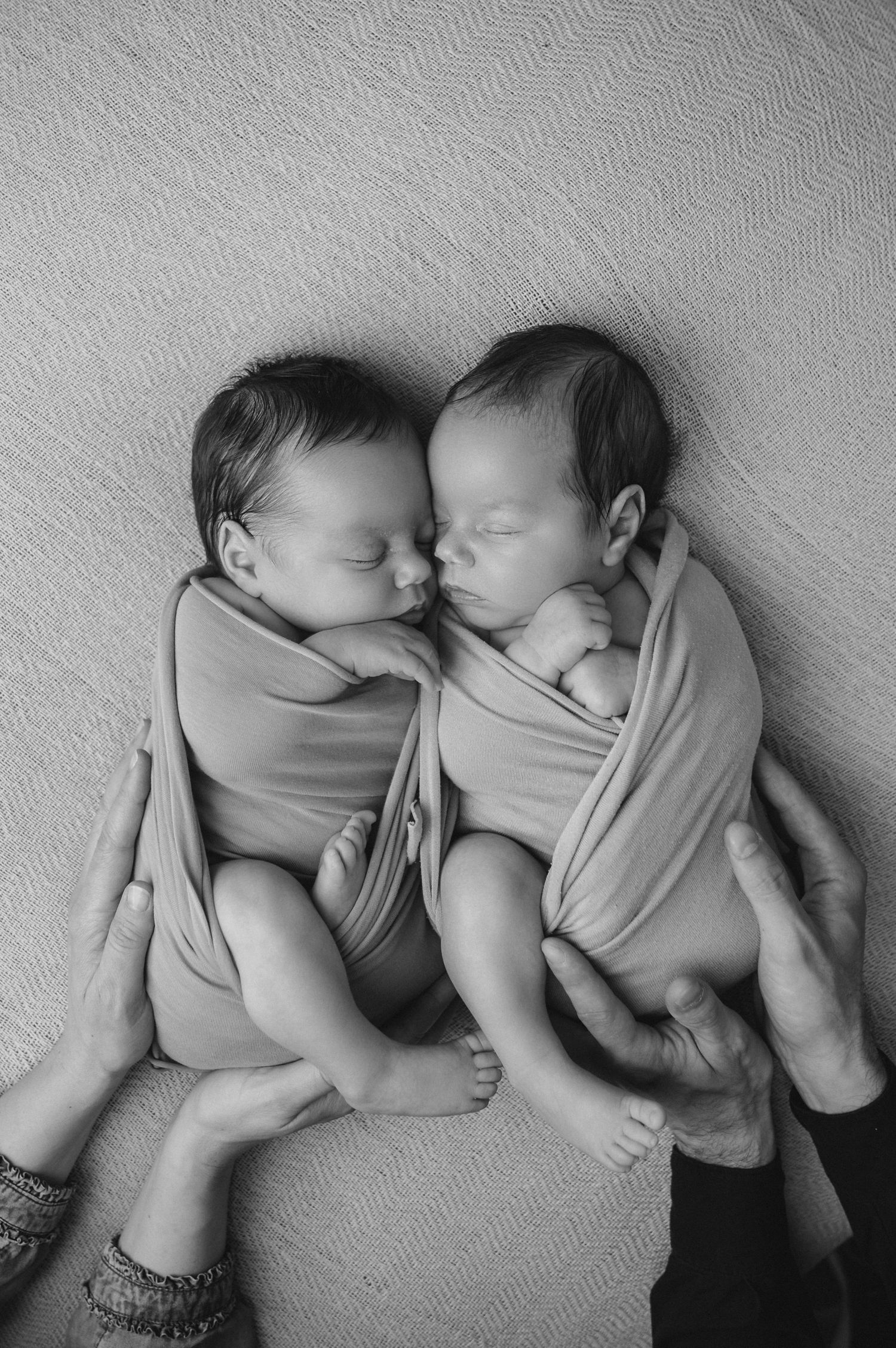 Two sleeping newborn babies wrapped in beige fabric lying next to each other in black and white portrait.