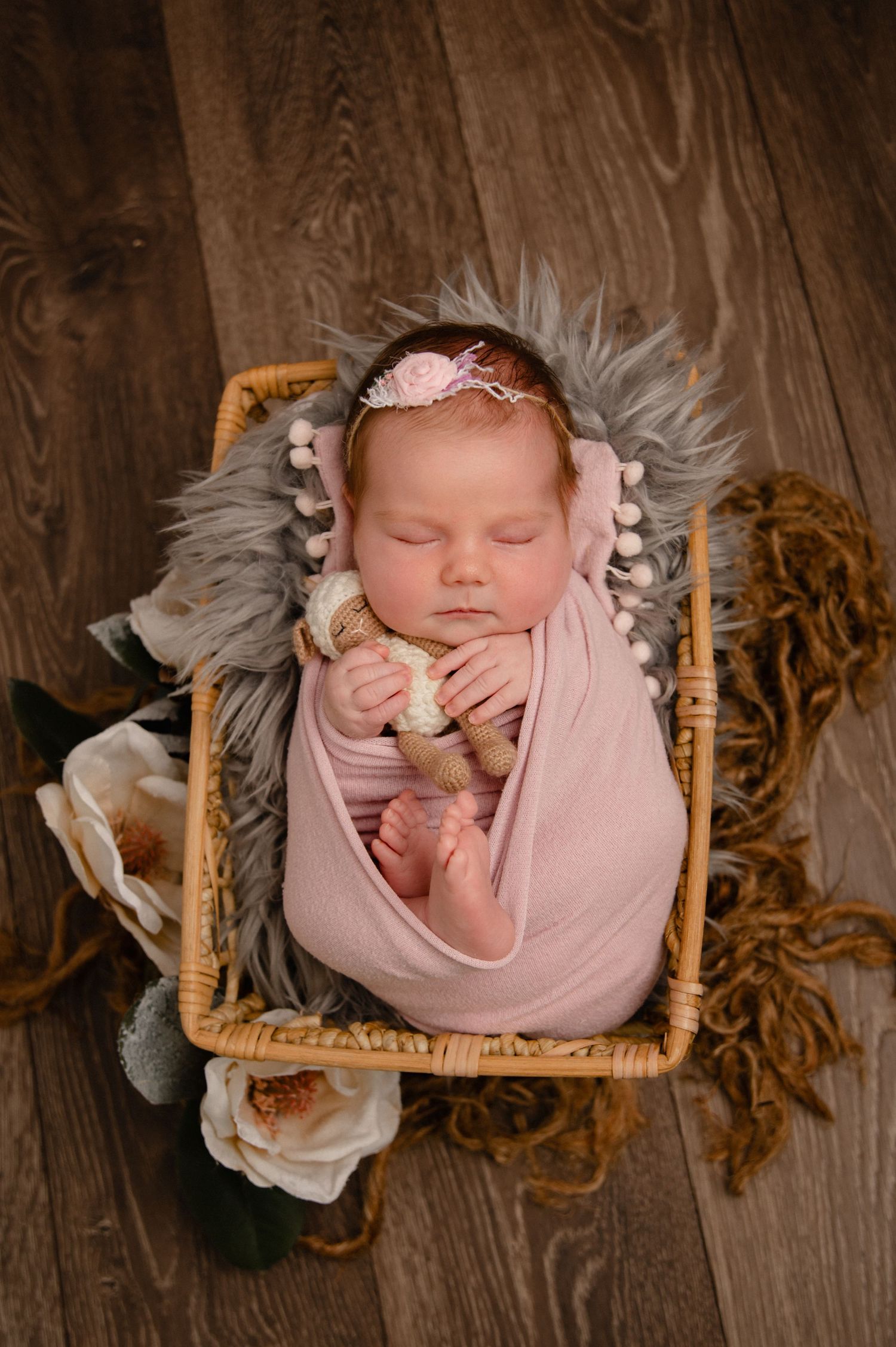 Sleeping newborn wrapped in pink swaddle with headband resting in fur-lined basket with floral accents.