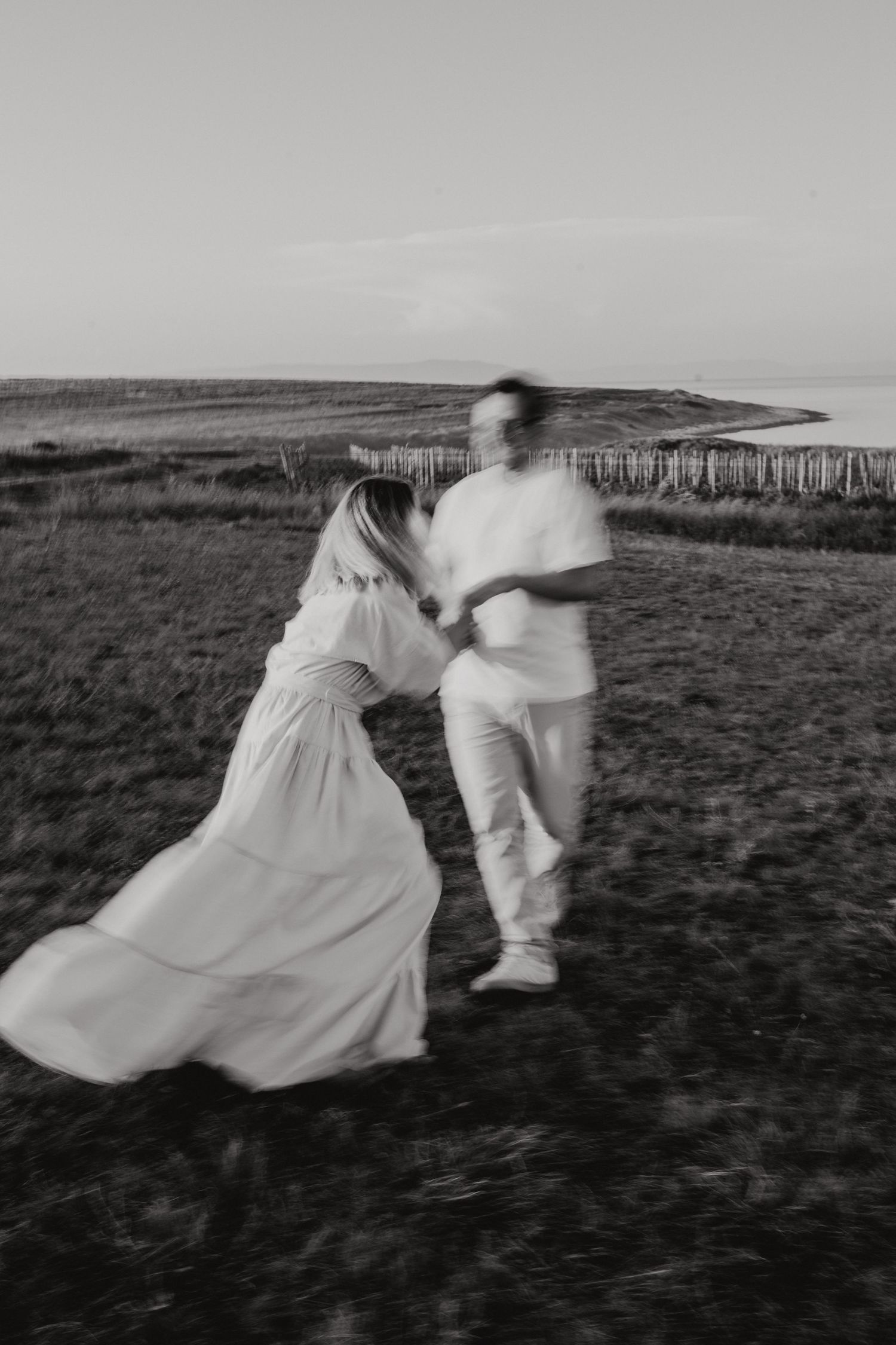 Two figures in white dance across a moody black and white landscape with rolling hills in the distance.