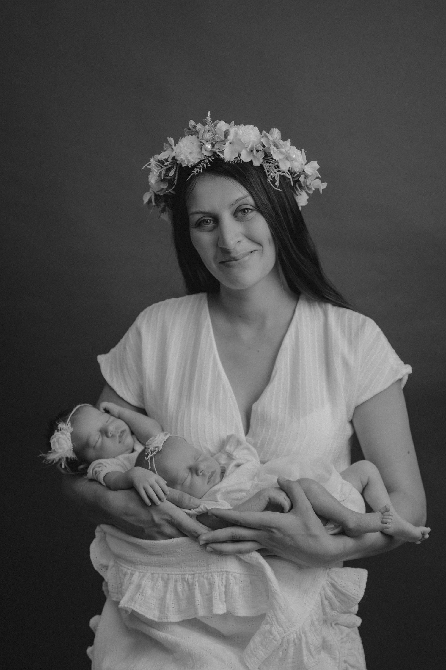 Mother in white dress and floral crown holds sleeping twins in black and white portrait.