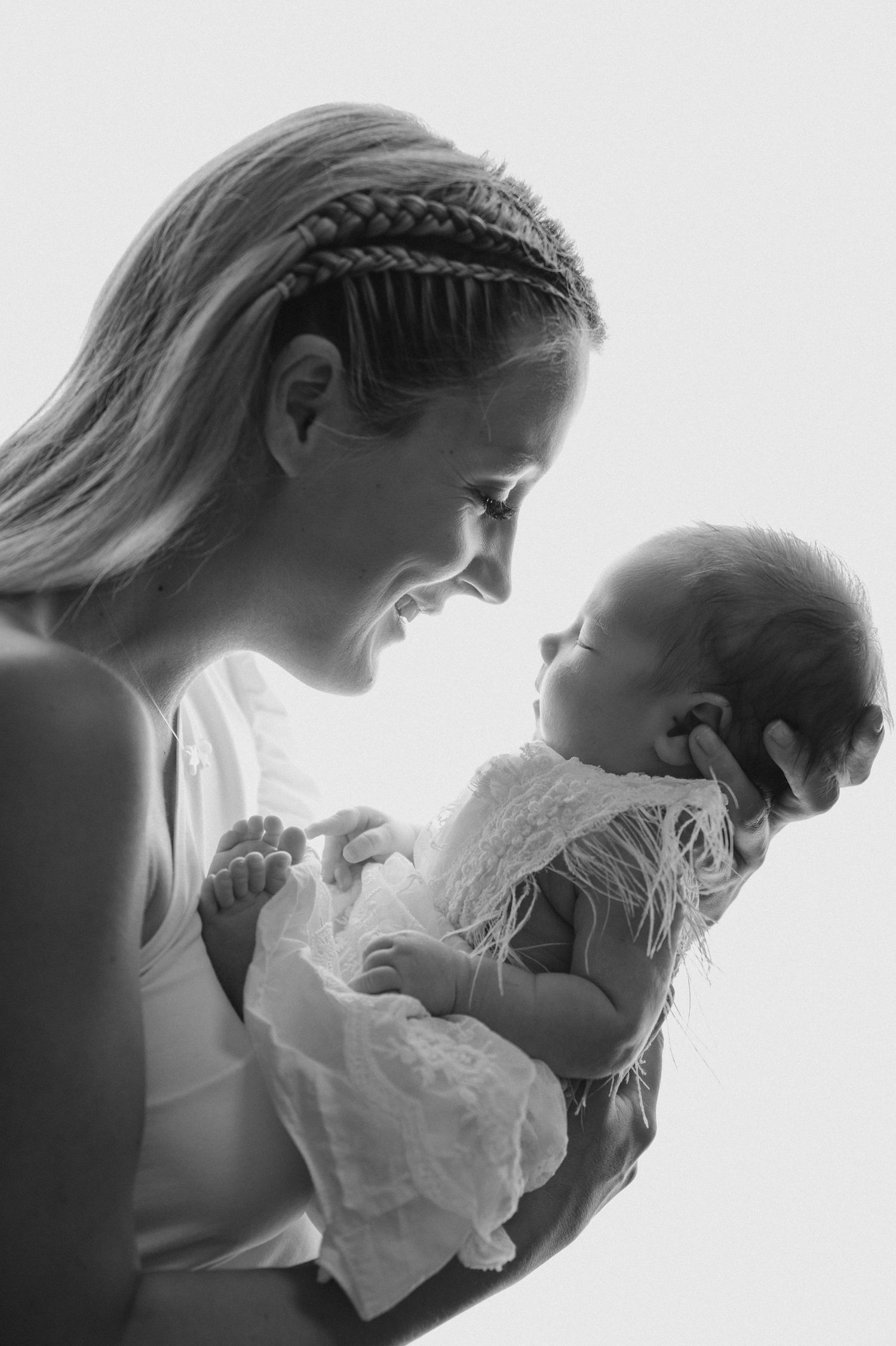 Side profile of mother with braided hair gazing at baby in white lace dress.