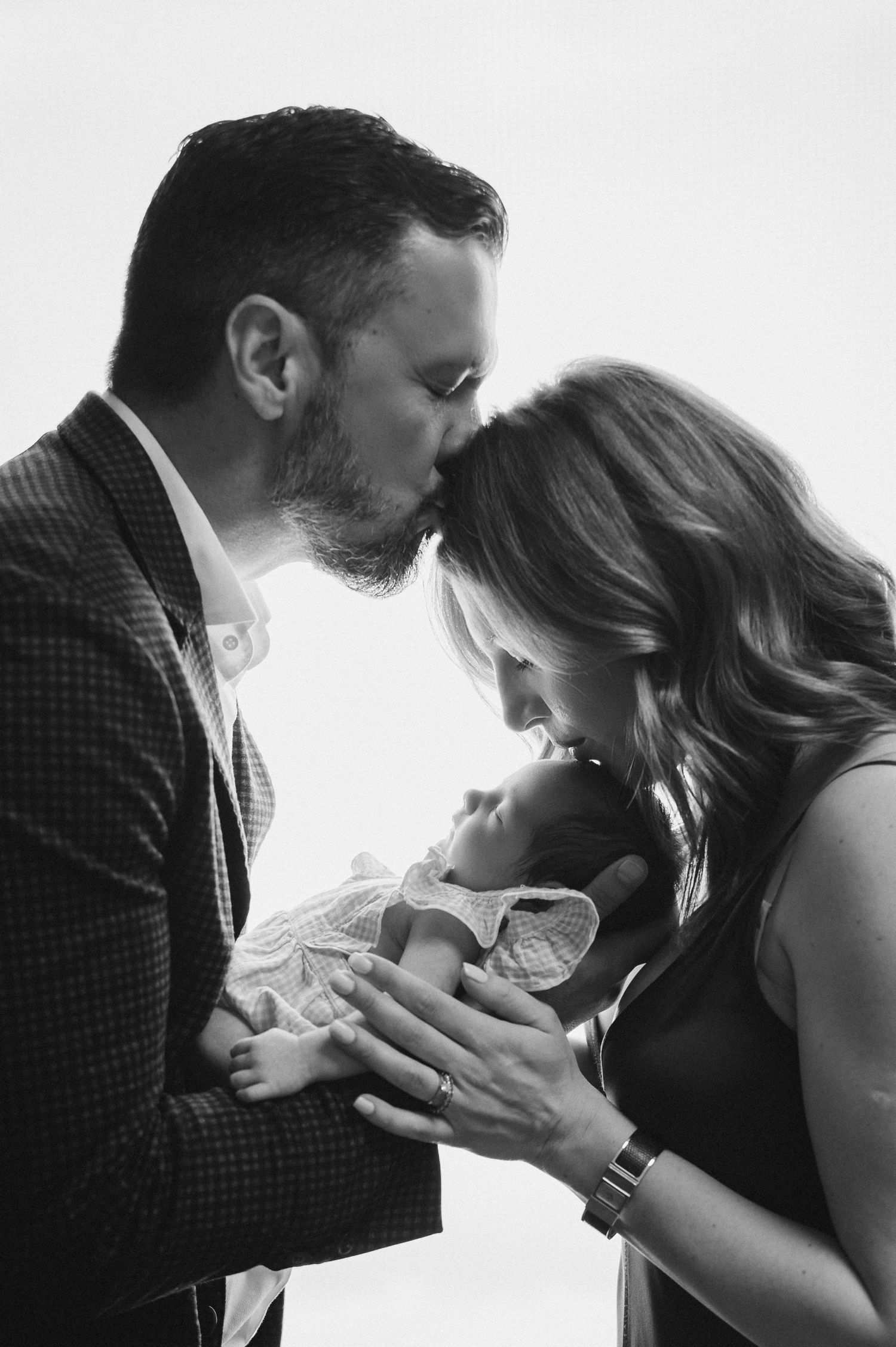 A tender black and white family portrait showing parents lovingly embracing their newborn baby together.