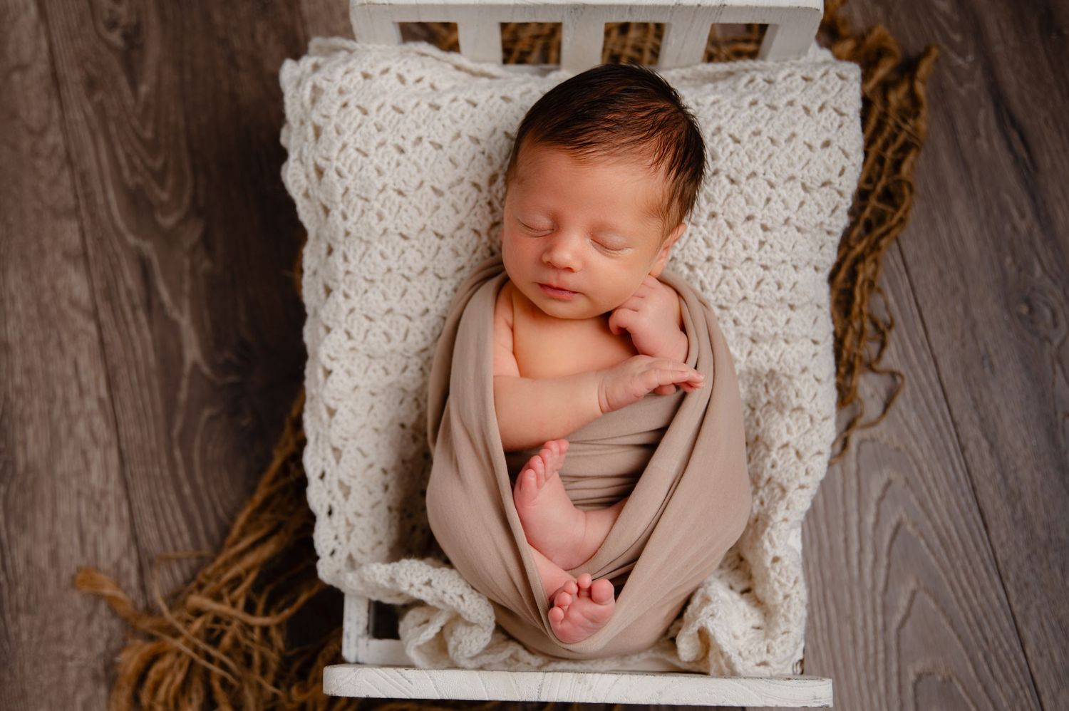 Sleeping newborn wrapped in tan swaddle on white crochet blanket in rustic setting.