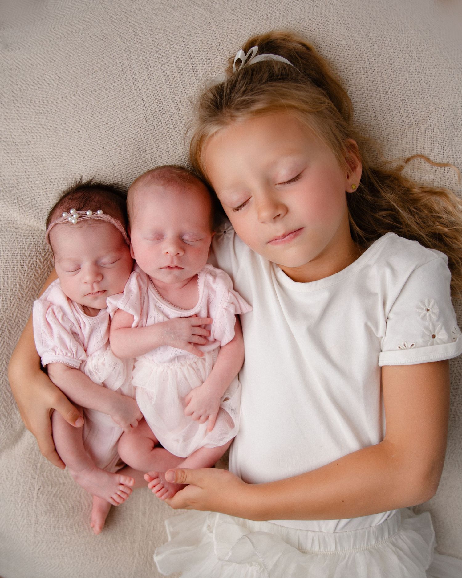 Young girl lying next to twin newborn babies on white fabric.