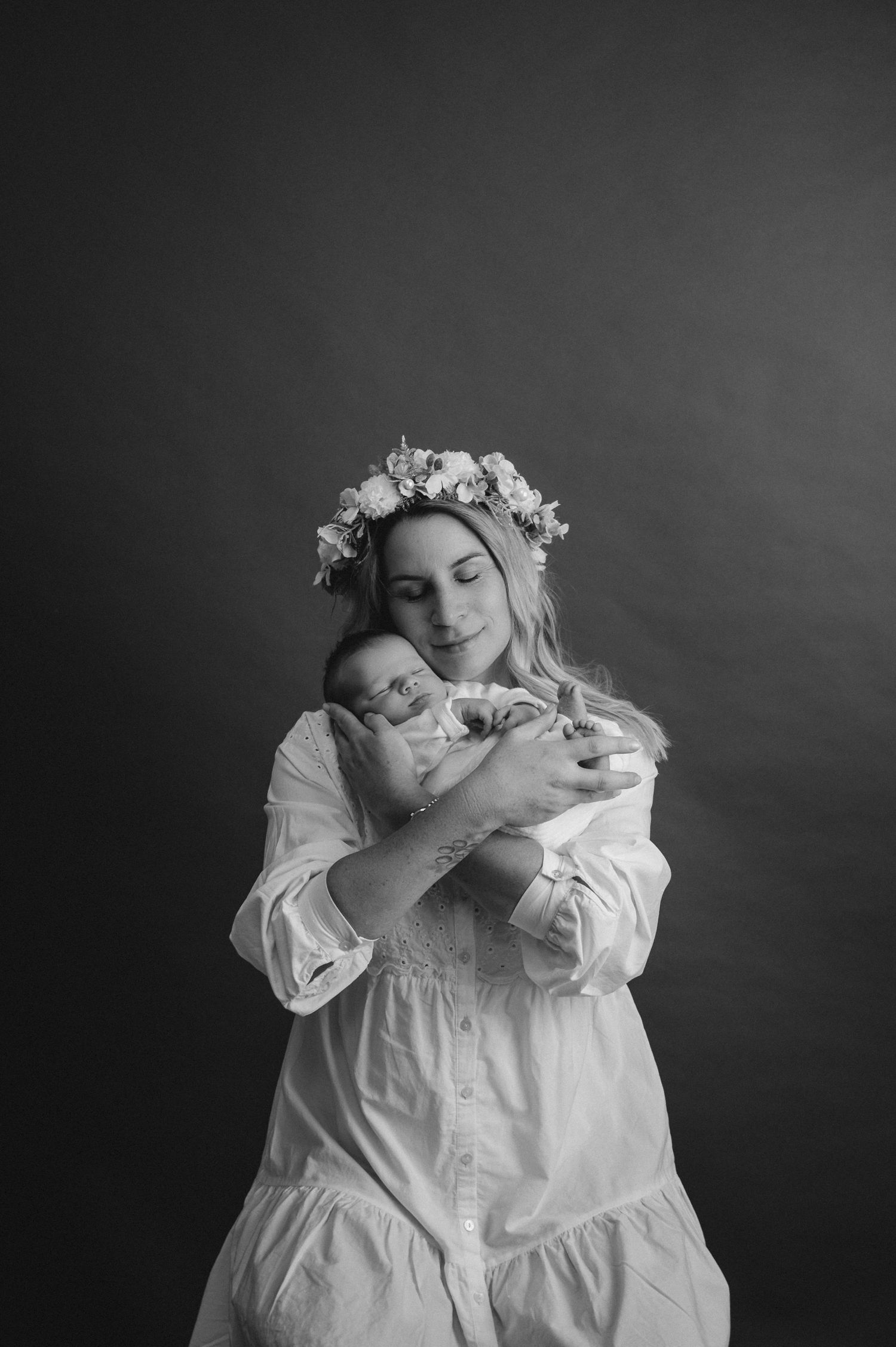 Black and white portrait of mother in white dress with flower crown holding sleeping newborn.