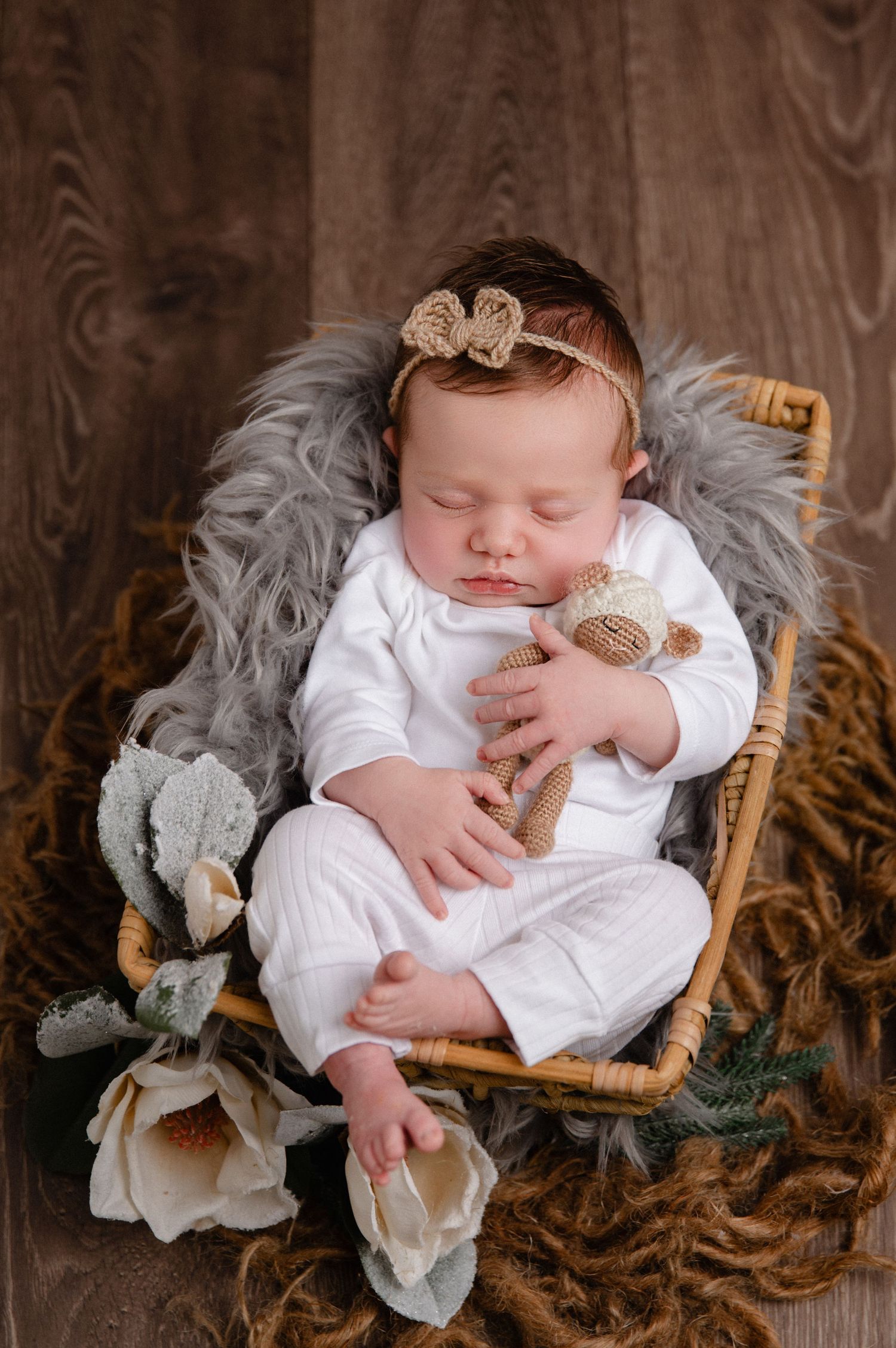 Sleeping baby in white outfit with bow headband resting in basket with grey fur and flowers.
