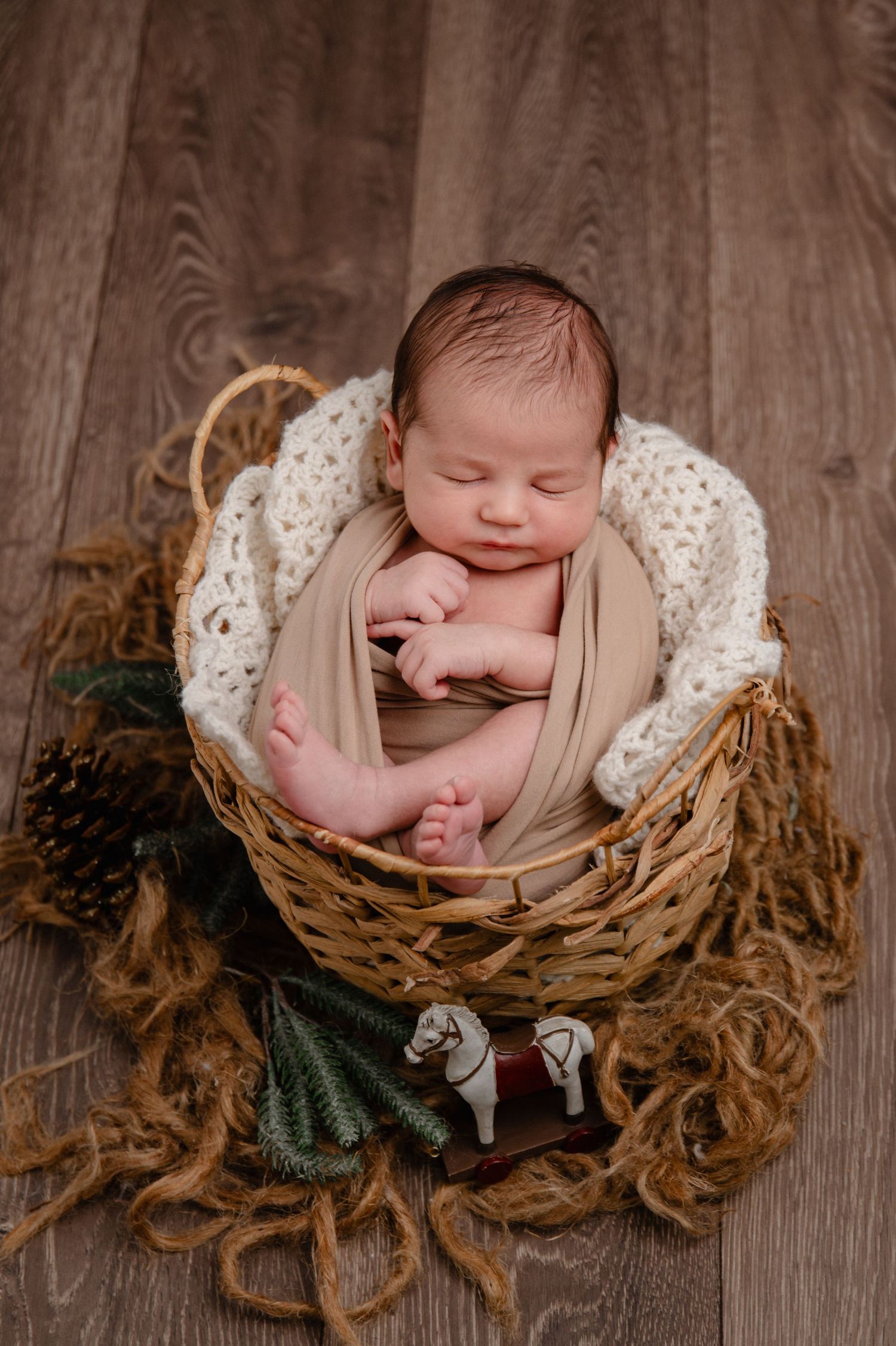 Sleeping newborn wrapped in beige fabric nestled in a rustic basket with pine cones.