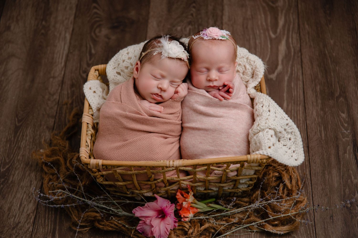 Twin newborns sleeping peacefully in a wicker basket wearing matching pink wraps and white headbands.