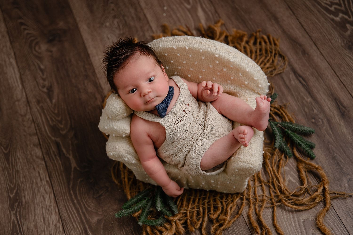 Alert newborn lying in cozy cream-colored basket with greenery on wooden surface.