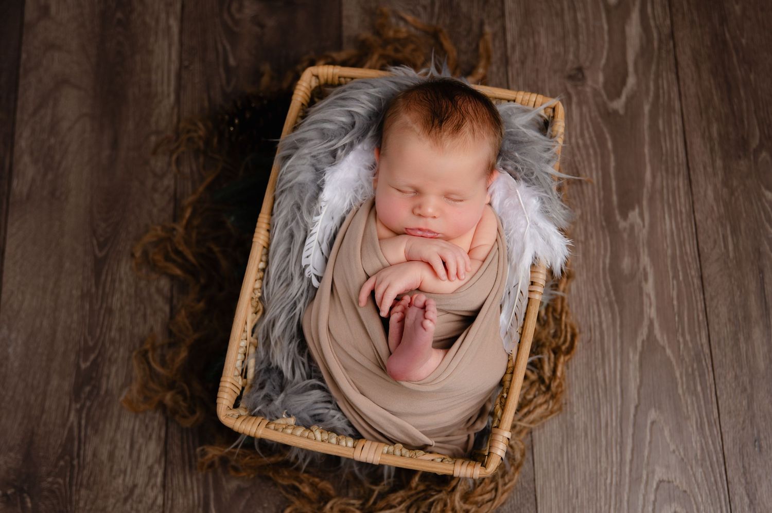 Sleeping newborn wrapped in beige fabric nestled in a basket with gray fur lining on wooden floor.