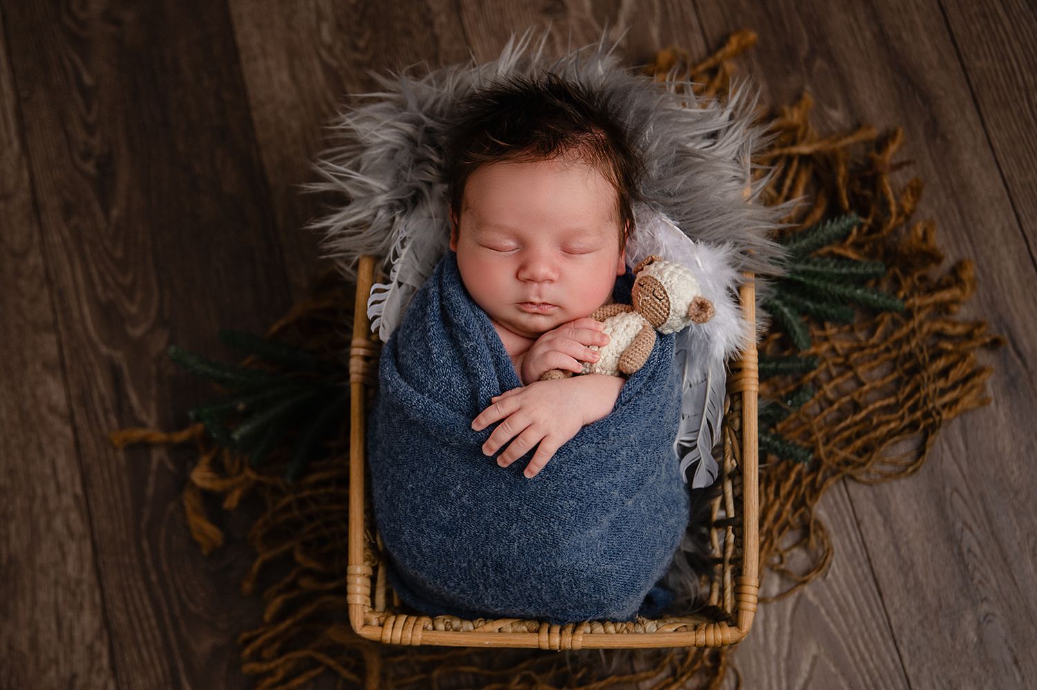Sleeping newborn wrapped in blue blanket nestled in rustic basket with foliage accents on wooden floor.