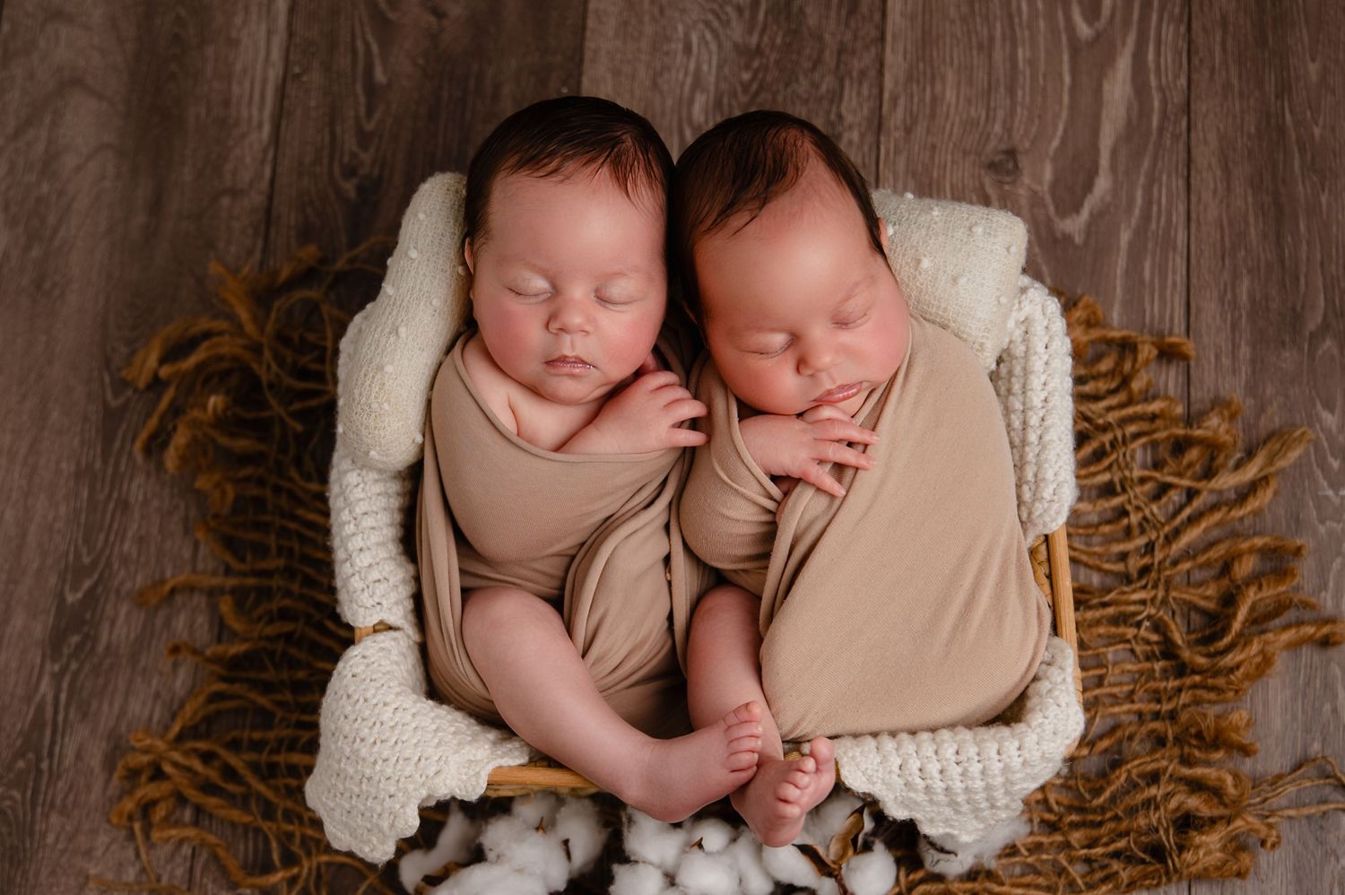 Two swaddled newborns sleeping side by side in a cozy white blanket-lined wicker basket on a wooden floor.