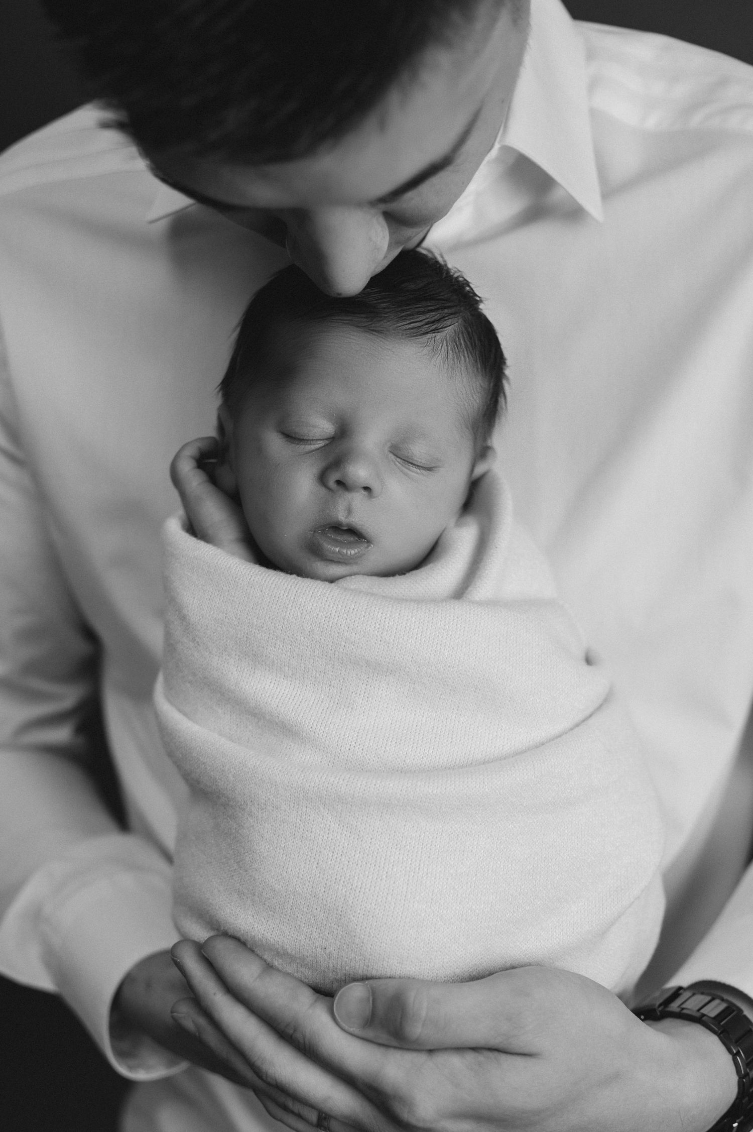 Intimate black and white portrait of father kissing newborn's head.