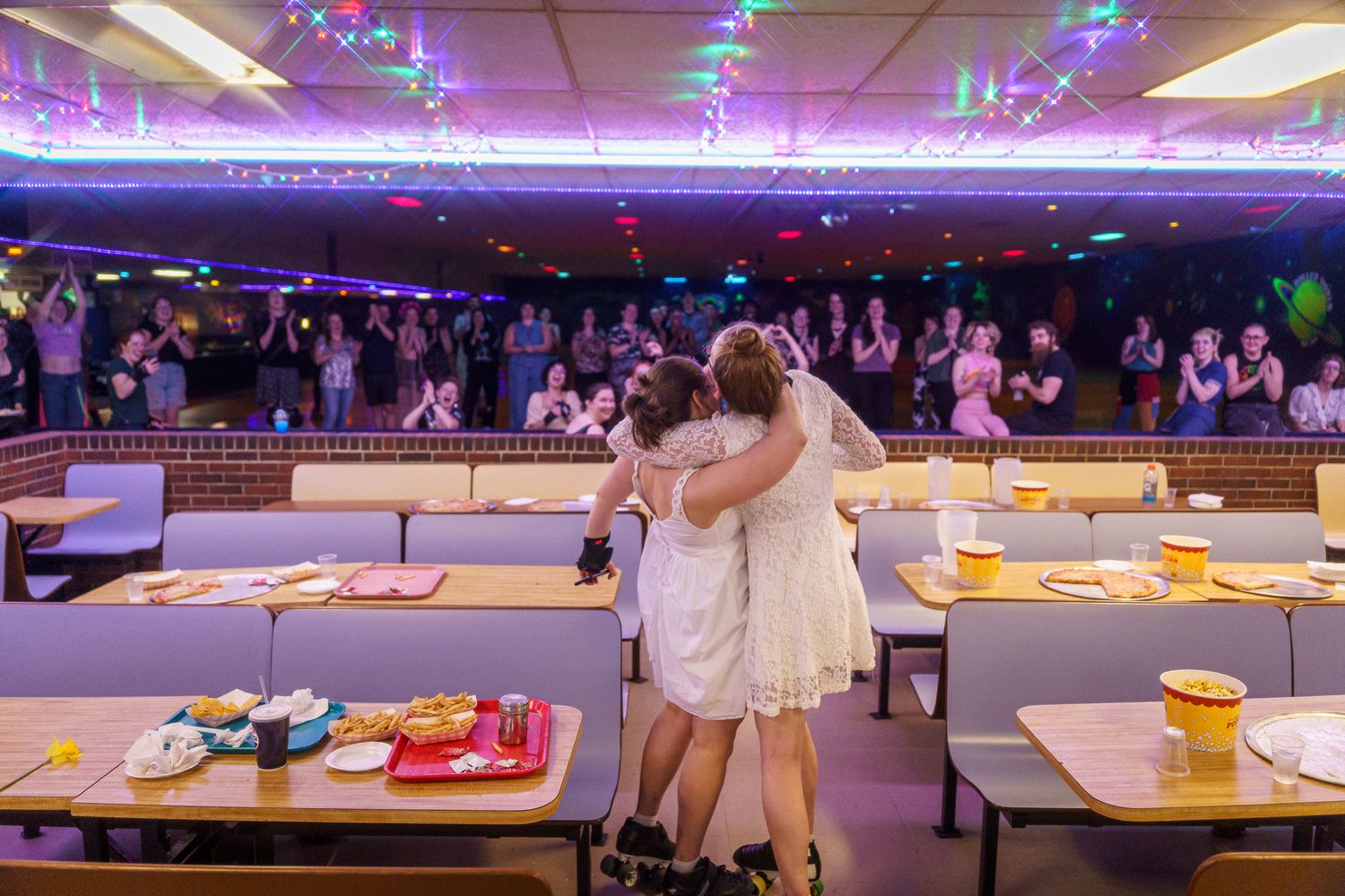 Two people embrace while dancing inside a retro-style bowling alley with neon lights and tables set for dining.