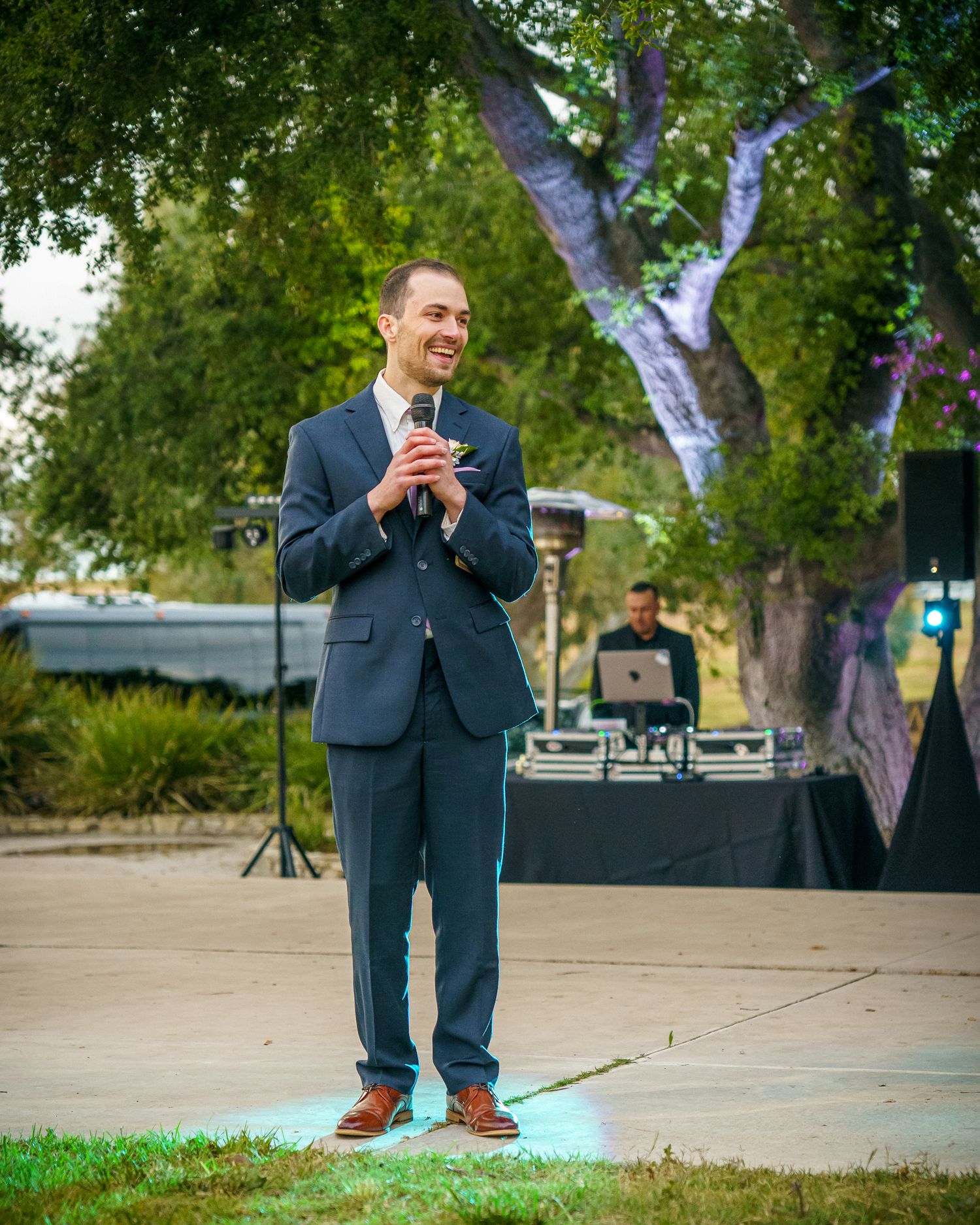 A speaker in a navy suit holds a microphone while presenting outdoors with a DJ booth and trees in the background.