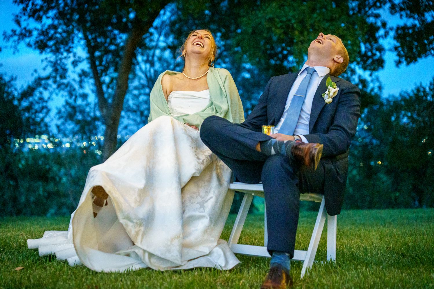 A bride and groom share a joyful candid moment while seated on white chairs on a lawn during their outdoor wedding.