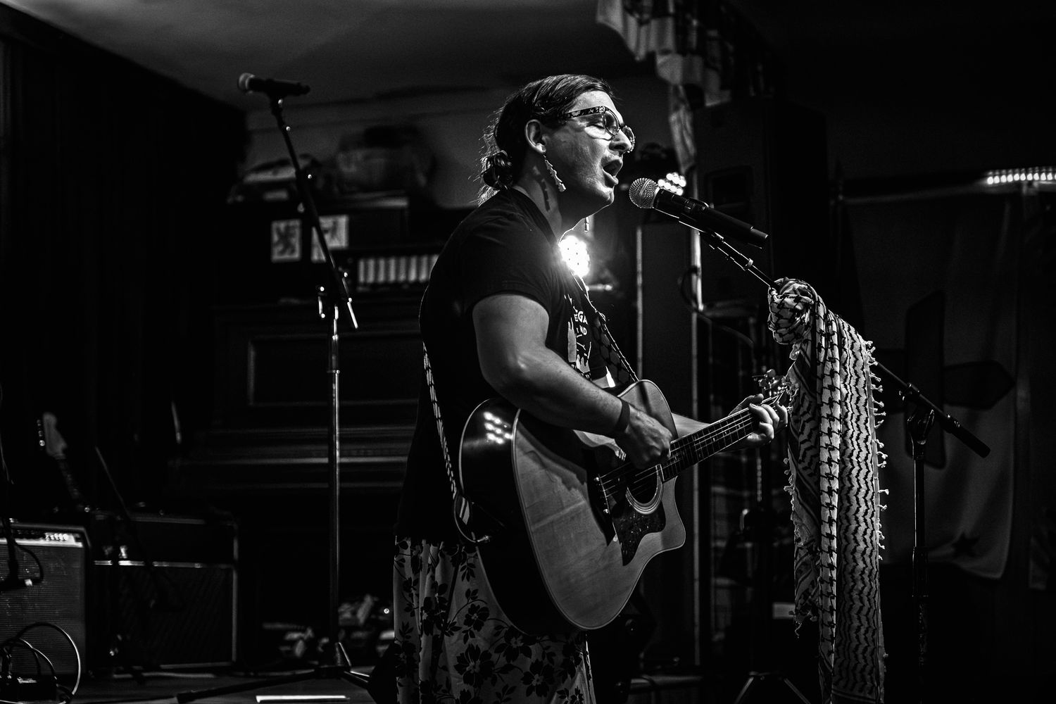 Performer in black shirt plays acoustic guitar on dark stage with dramatic lighting during live concert performance.