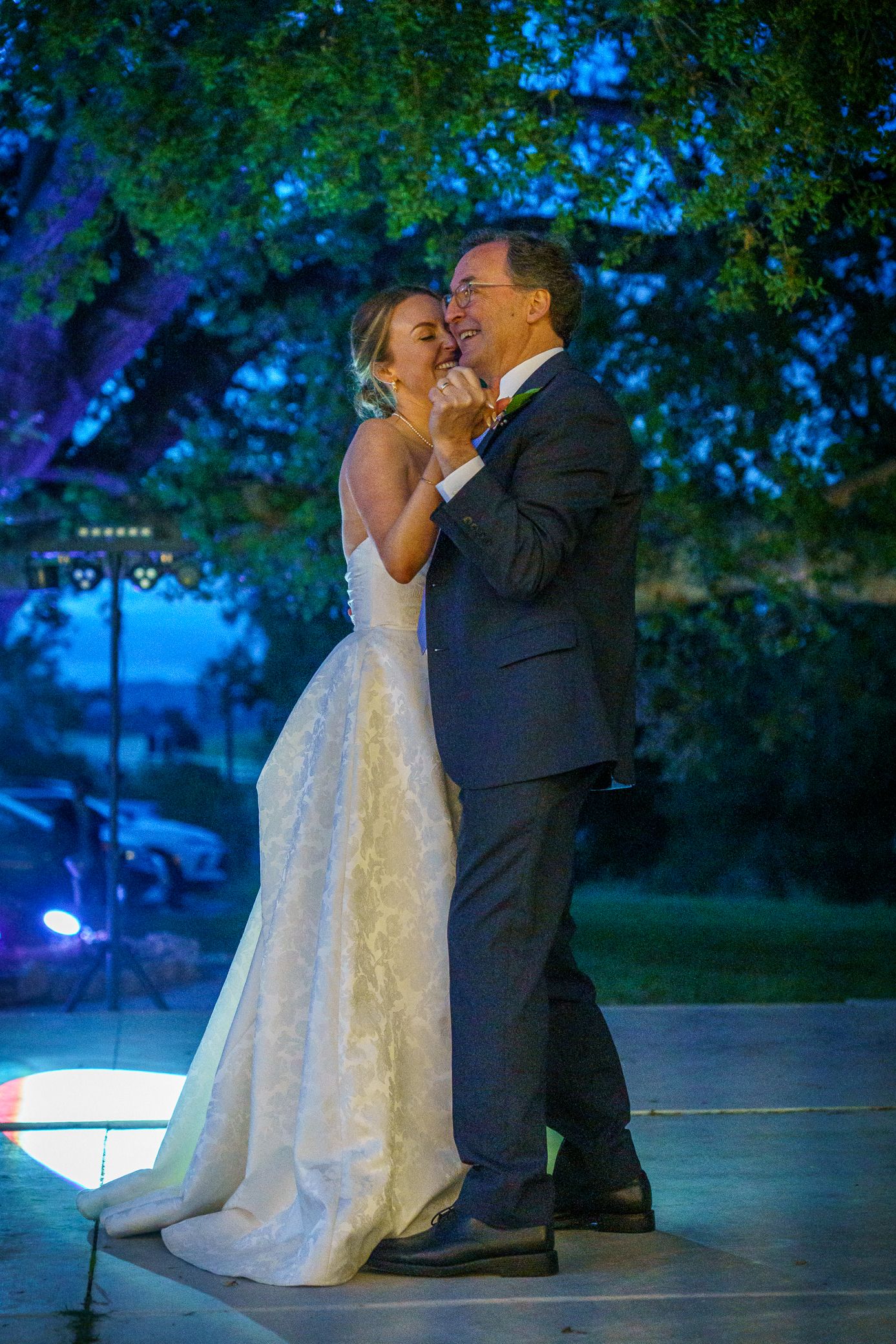 Two people in formal wedding attire share an intimate dance moment on an outdoor patio lit by blue evening light.