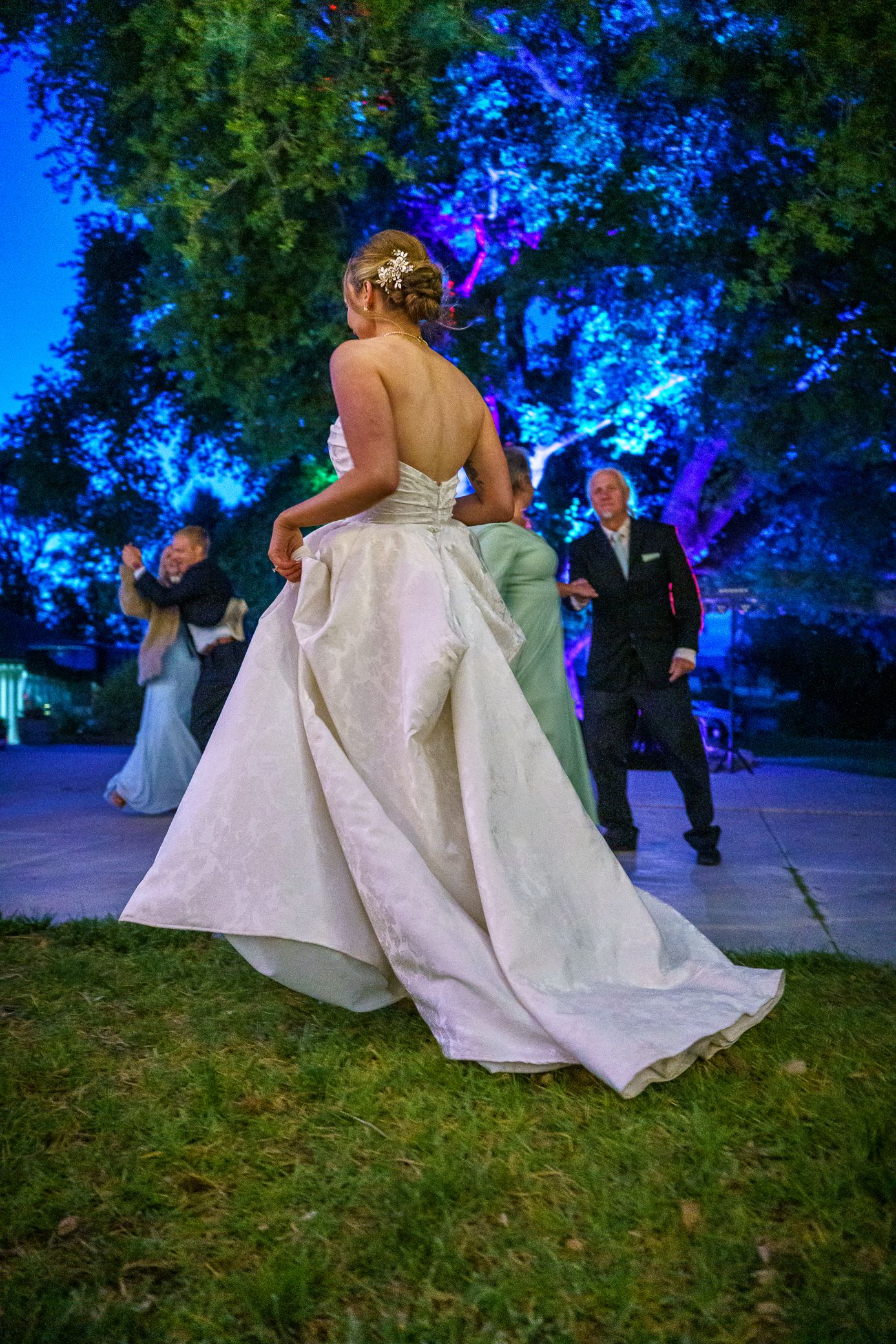 A bride in a flowing white wedding gown dances at an outdoor evening reception with trees illuminated by blue lights.