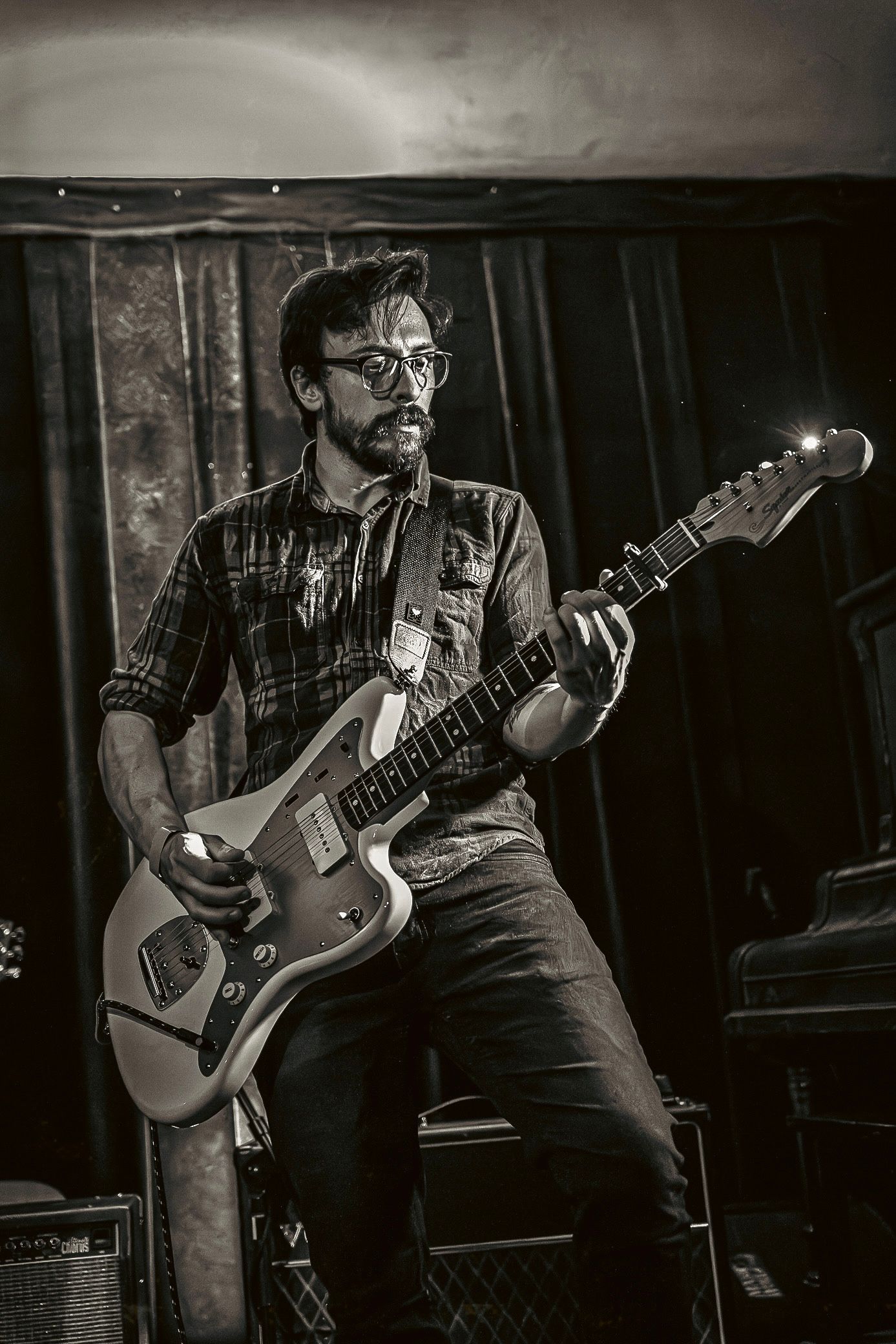 Black and white photo of a musician playing electric guitar on stage wearing plaid shirt and glasses.