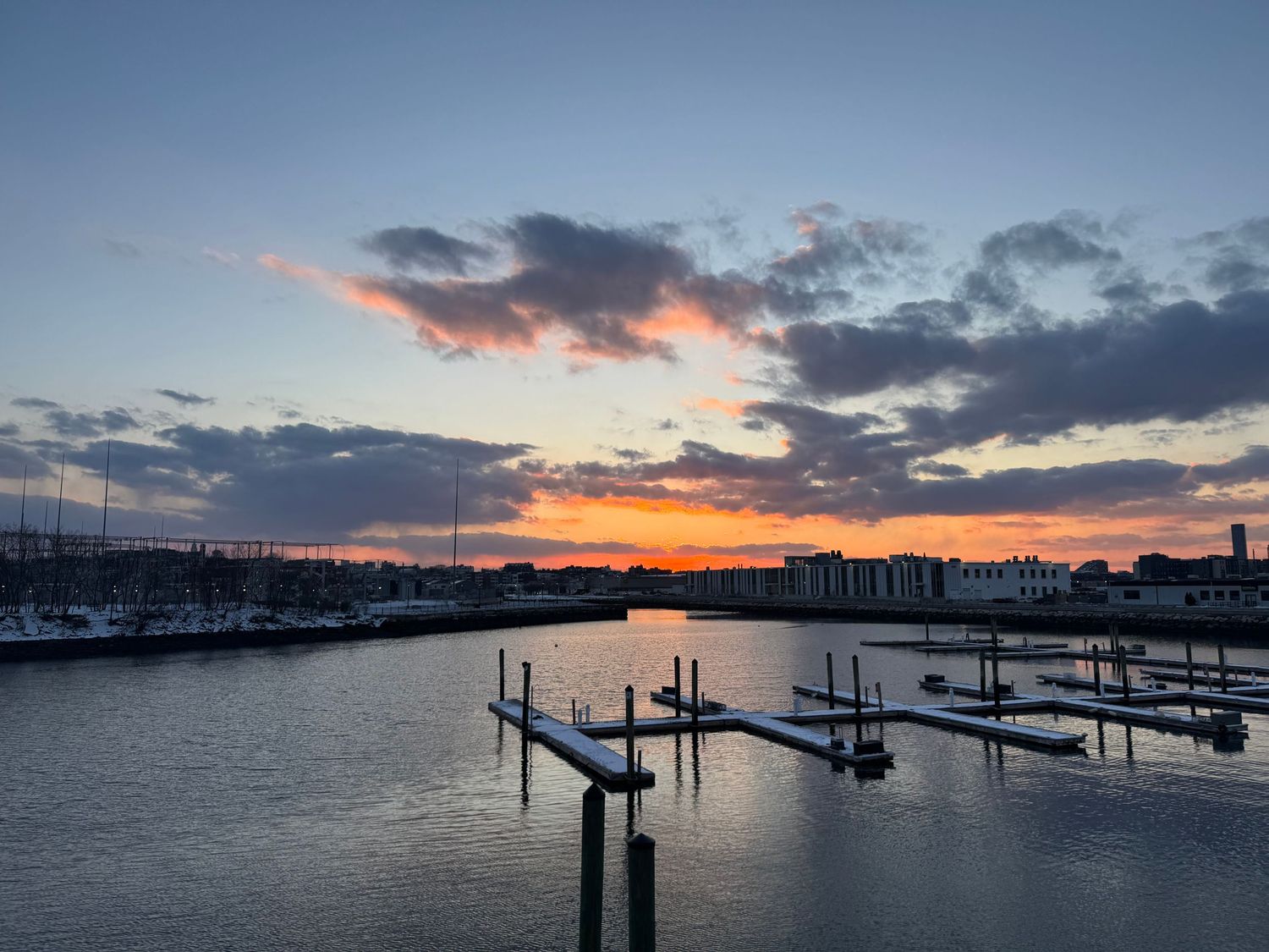 Boats docked at marina silhouetted against vibrant sunset sky with orange clouds reflecting on calm harbor waters.