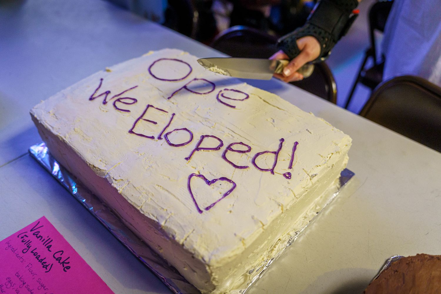 A simple white frosted cake with purple handwritten text reading 'Ope We Eloped!' and a heart decoration.