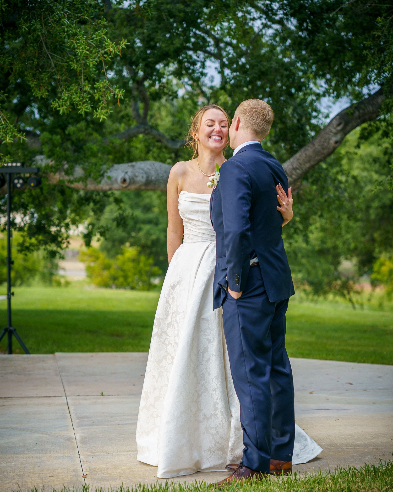 A couple in wedding attire share a tender moment outdoors with the bride in white and groom in navy blue suit.