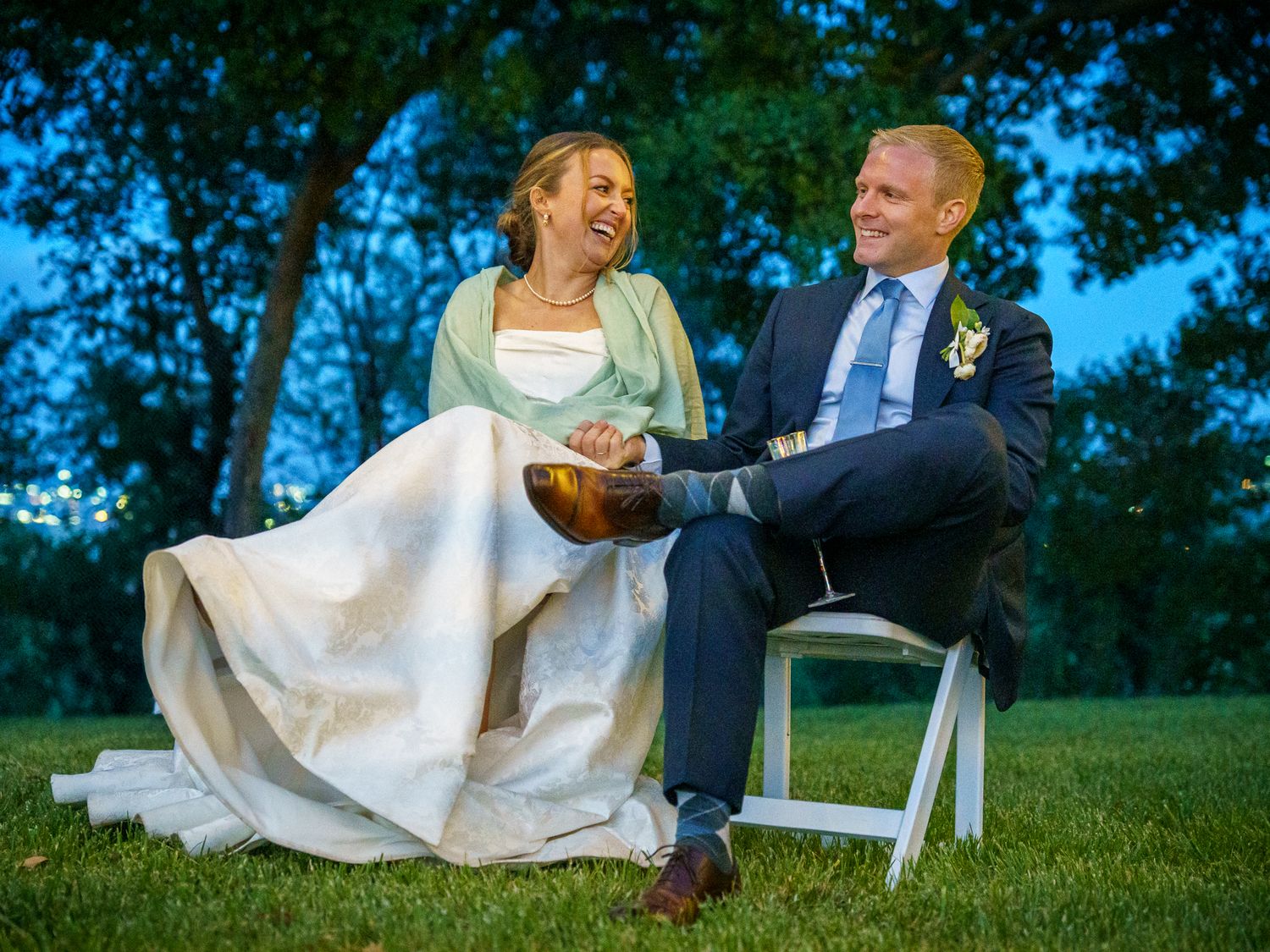 Two newlyweds share a joyful moment while seated on chairs outdoors during their outdoor evening wedding ceremony.