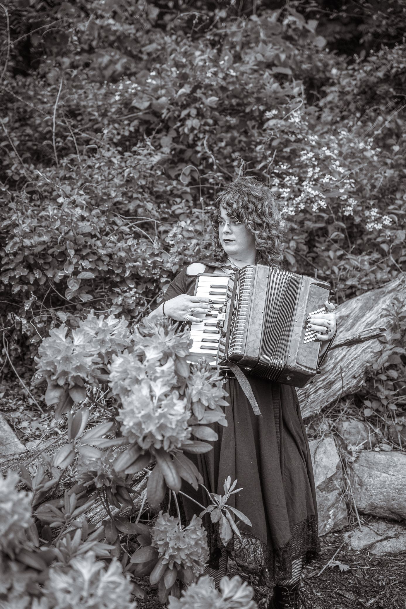 Black and white portrait of an accordion player in a dress standing among lush garden foliage.