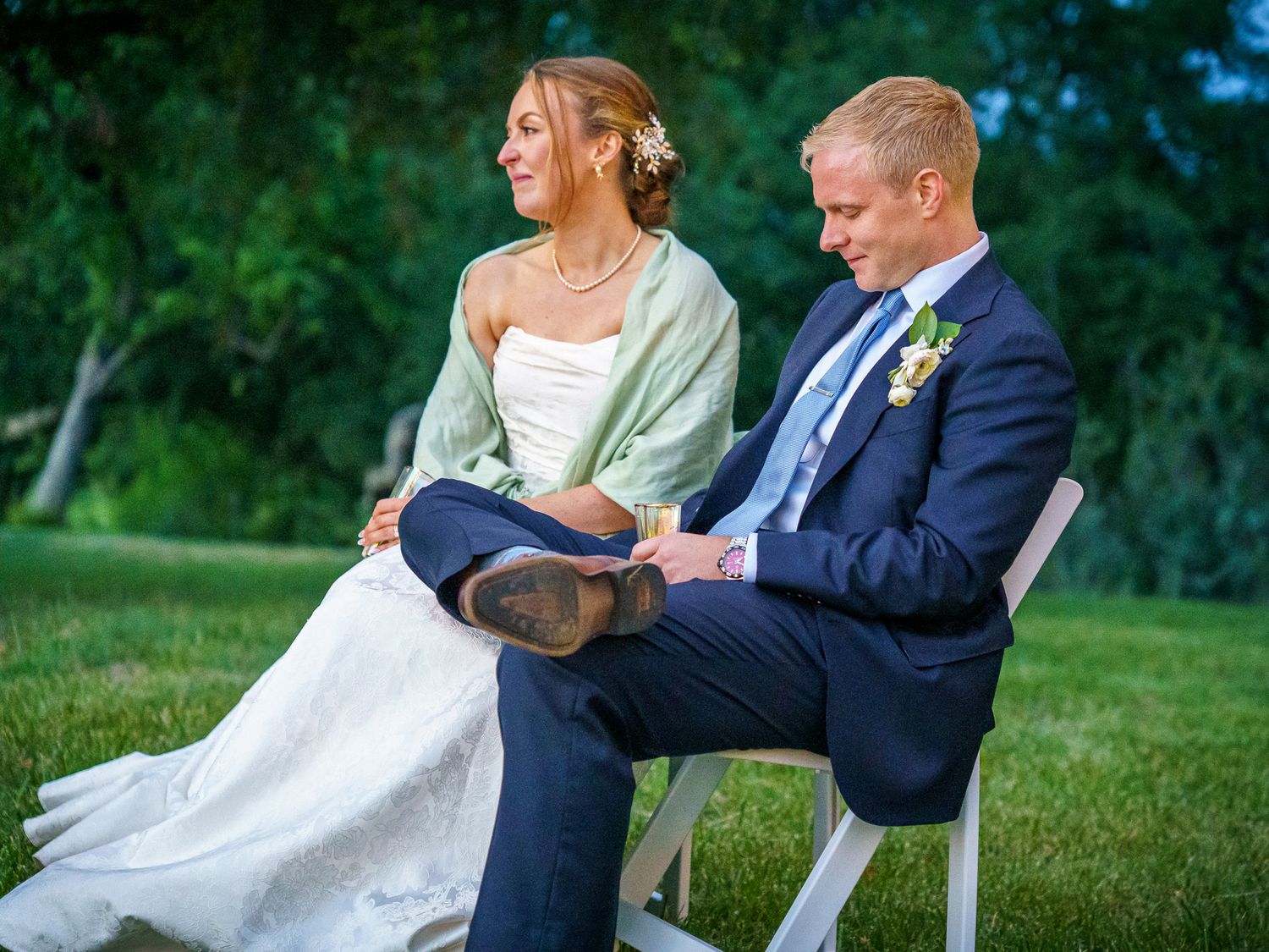 A wedding couple sits casually on white chairs outdoors in a garden setting during their ceremony.