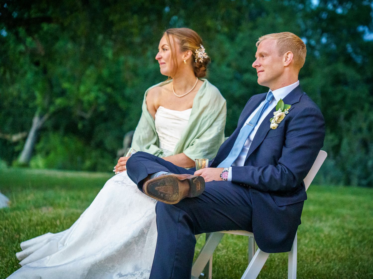 Couple dressed in wedding attire sits outdoors on chairs in a garden setting during golden hour.
