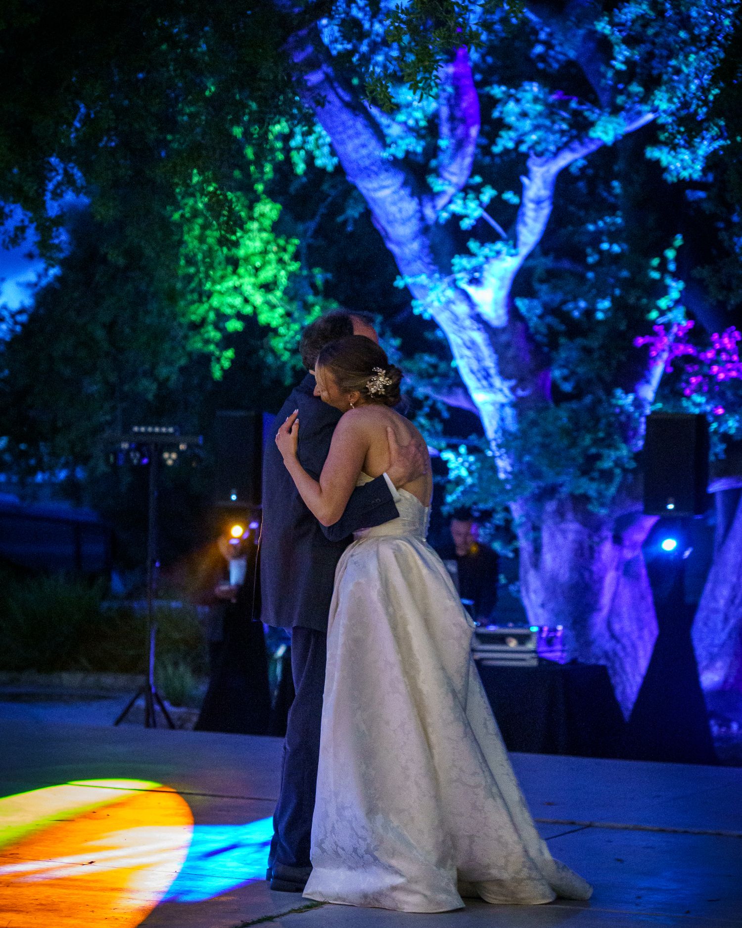 Couple dances at outdoor wedding reception under trees illuminated with blue, green and purple lighting effects.