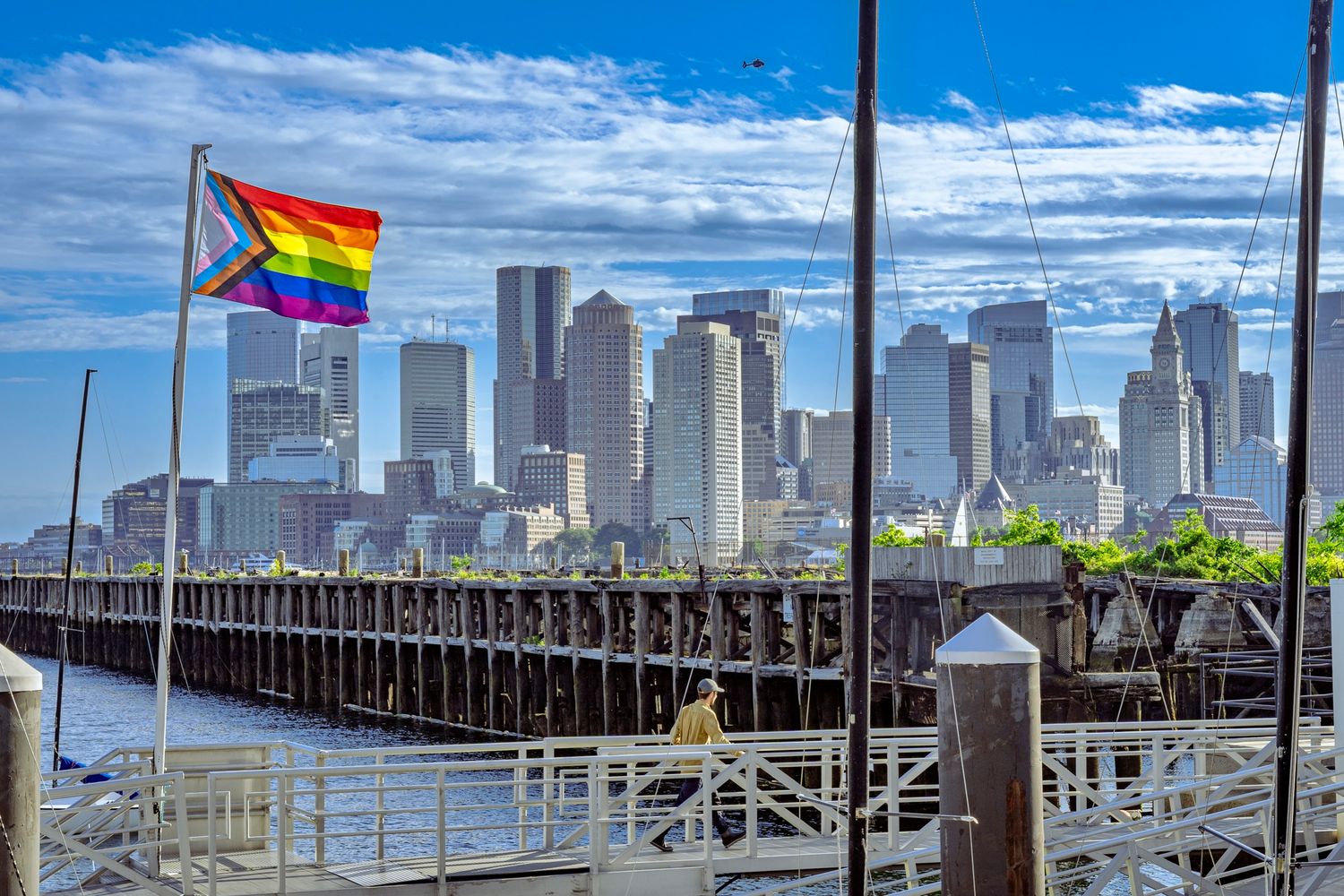 Pride flag waves over Boston skyline view from wooden pier with blue sky and clouds above city harbor.