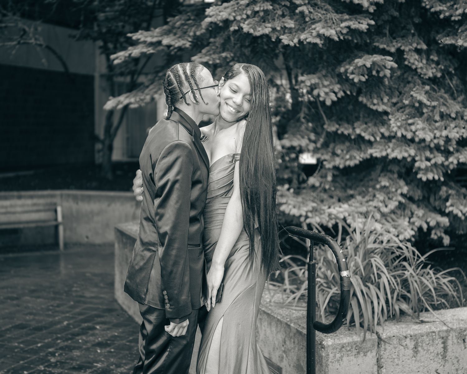 A black and white vintage photograph shows a romantic couple embracing outdoors near lush garden foliage.