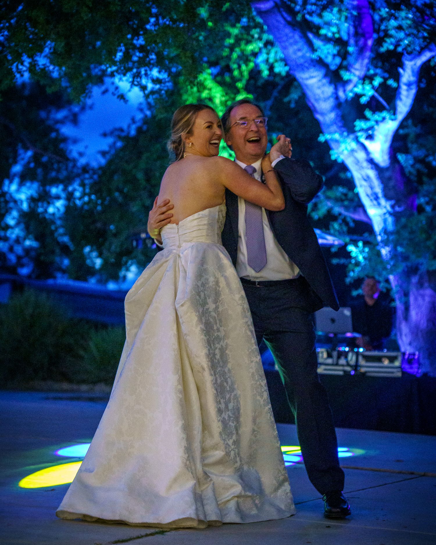 A couple shares a joyful dance moment at their wedding reception with blue and green illuminated trees in the background.