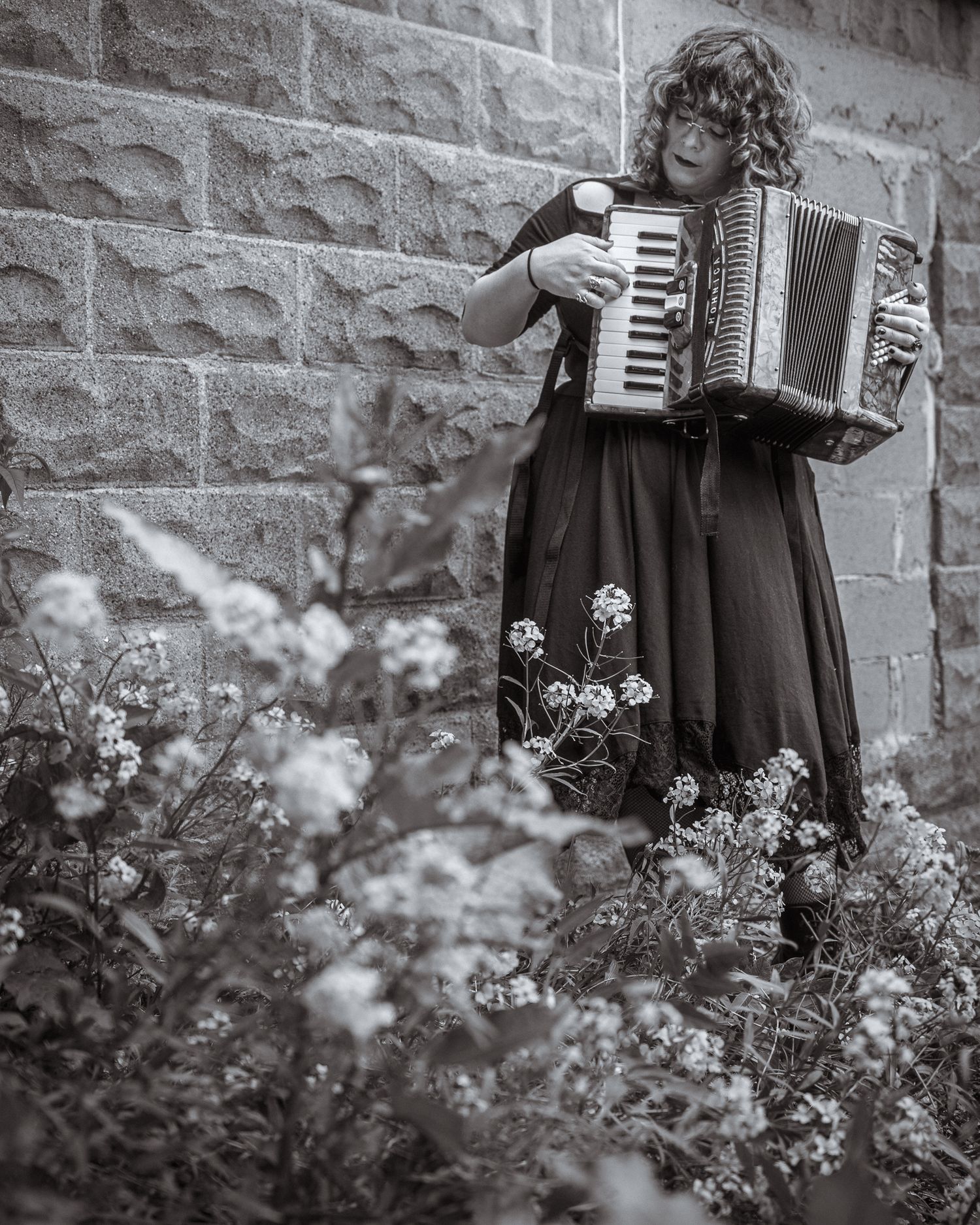 Person playing an accordion in a floral dress against stone wall with blooming flowers in black and white.