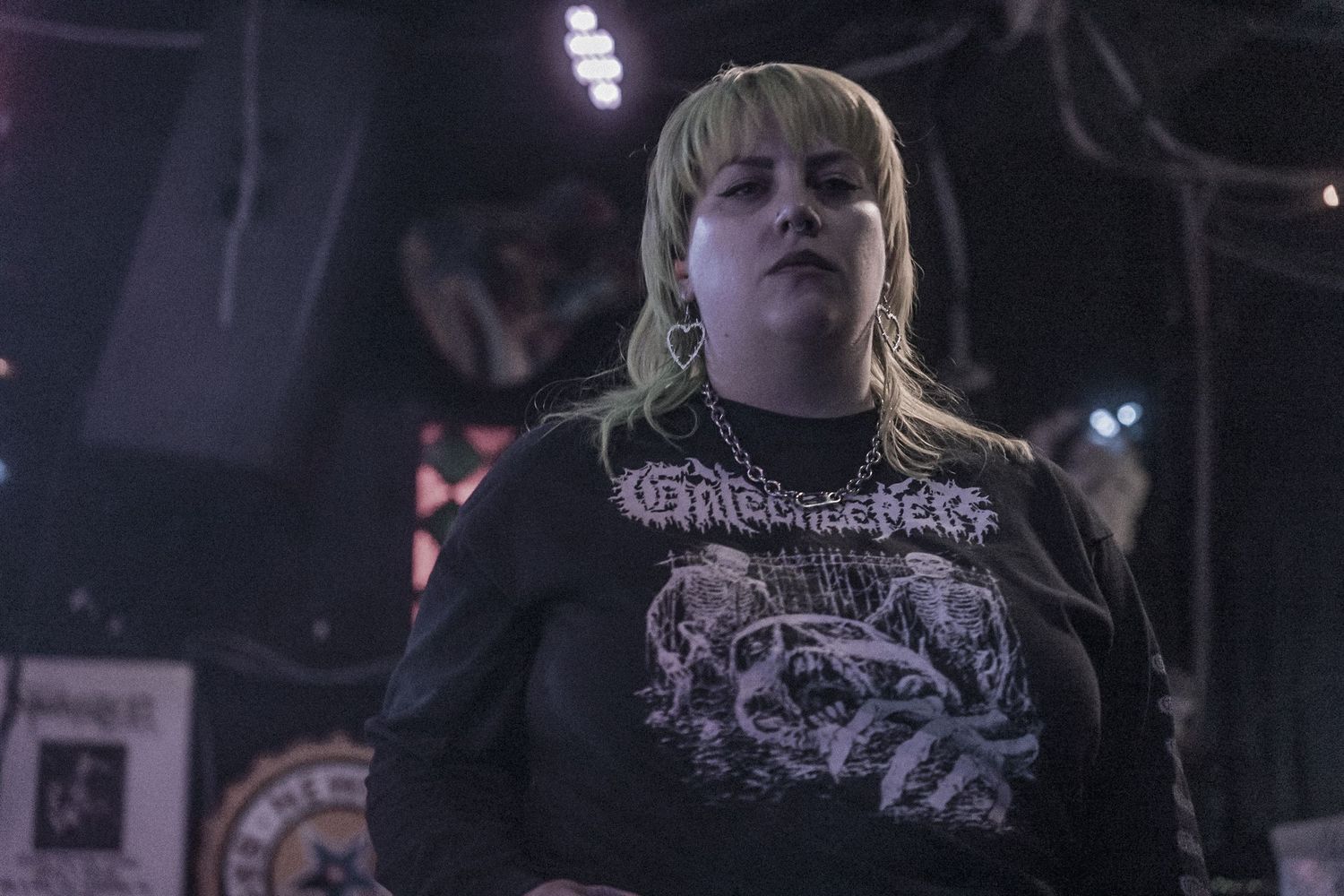 A concert scene featuring someone wearing a black metal band t-shirt in dimly lit atmospheric lighting.