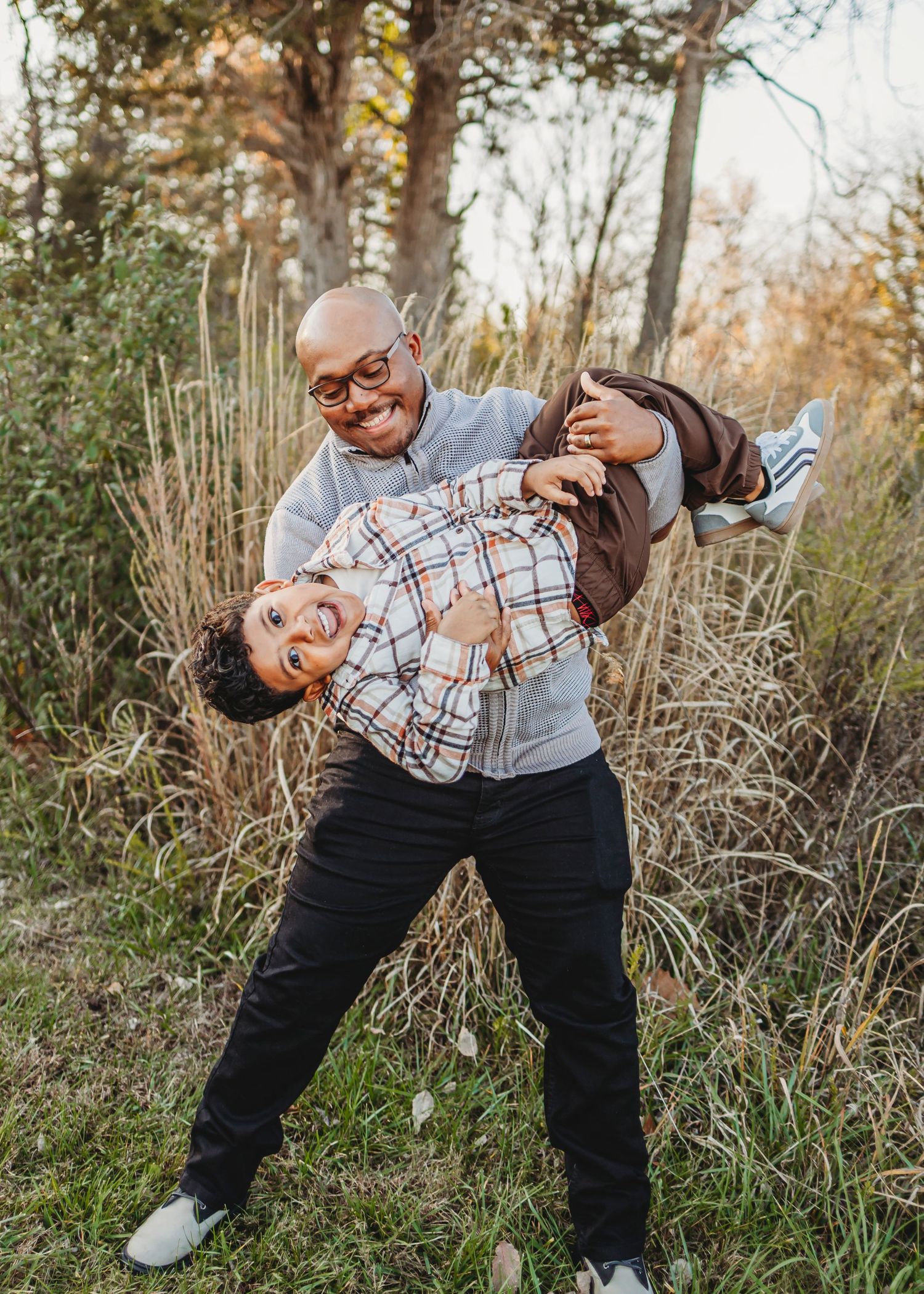 A person in a plaid shirt and dark pants playfully poses in tall dry grass during an autumn photoshoot.