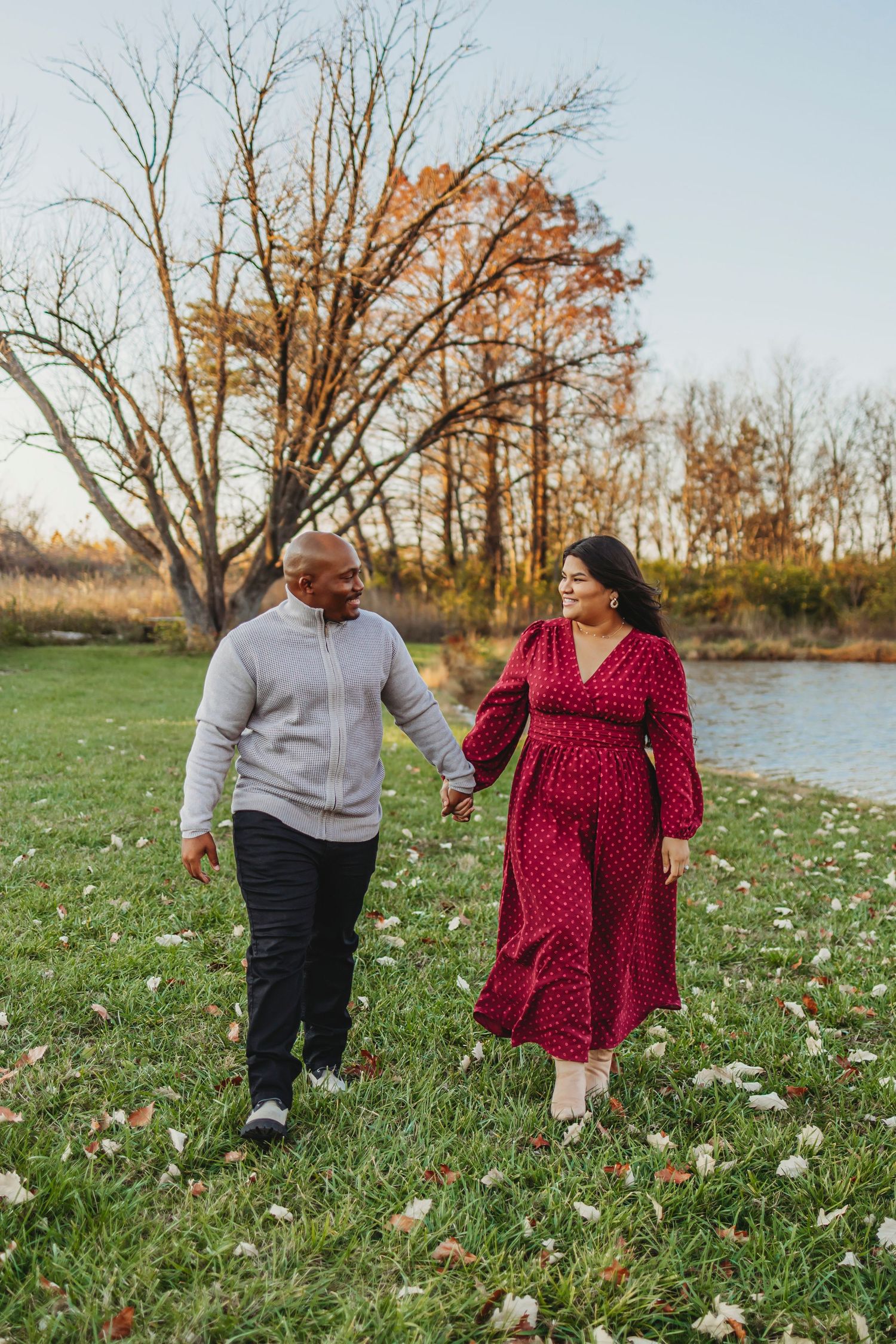 A couple strolls hand in hand through a grassy field near a pond during autumn with bare trees in the background.