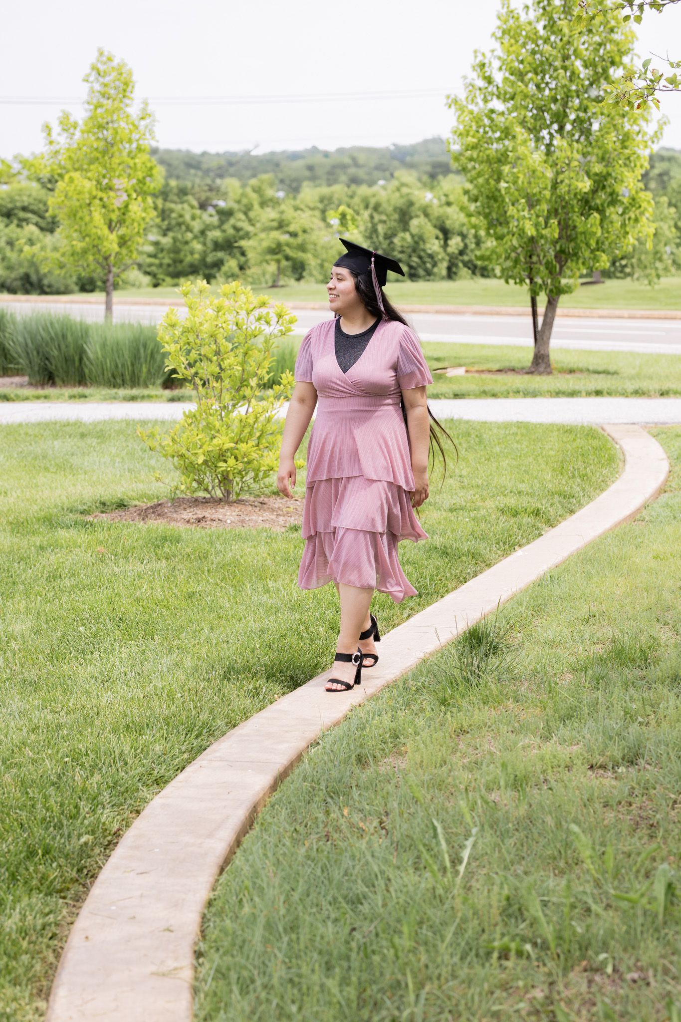A graduate in a flowing pink dress walks along a curved pathway surrounded by lush greenery.