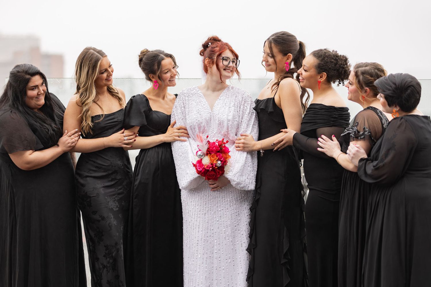 Wedding party in elegant black dresses gathering around the bride with red bouquet.
