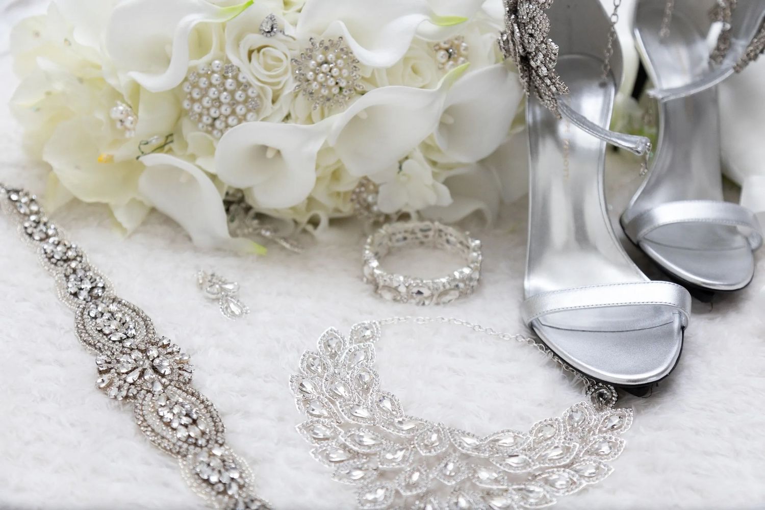 Wedding accessories including silver shoes, jewelry and white calla lilies arranged on white lace.