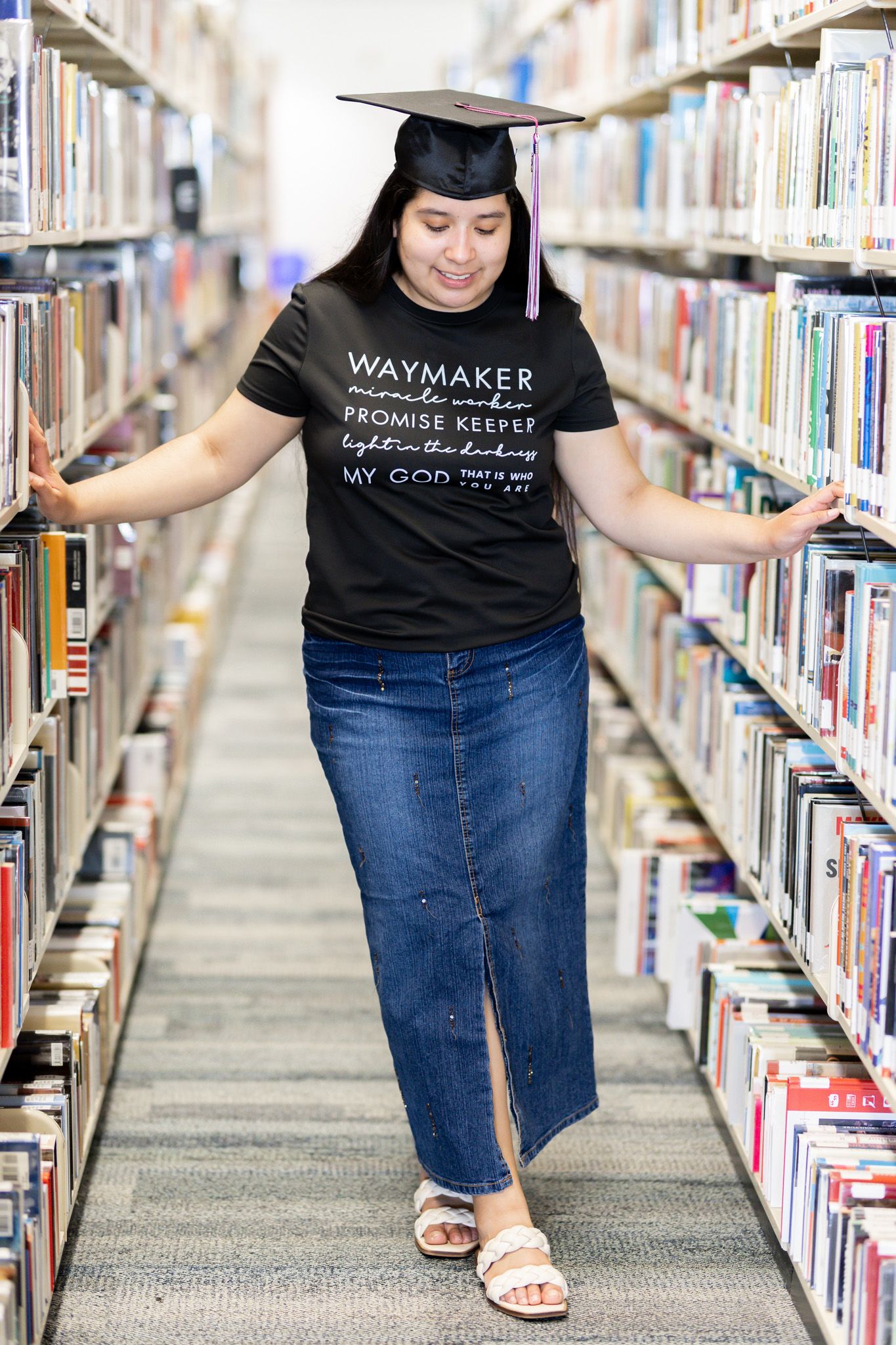 A graduate in cap and gown browses books in a library between tall shelves.